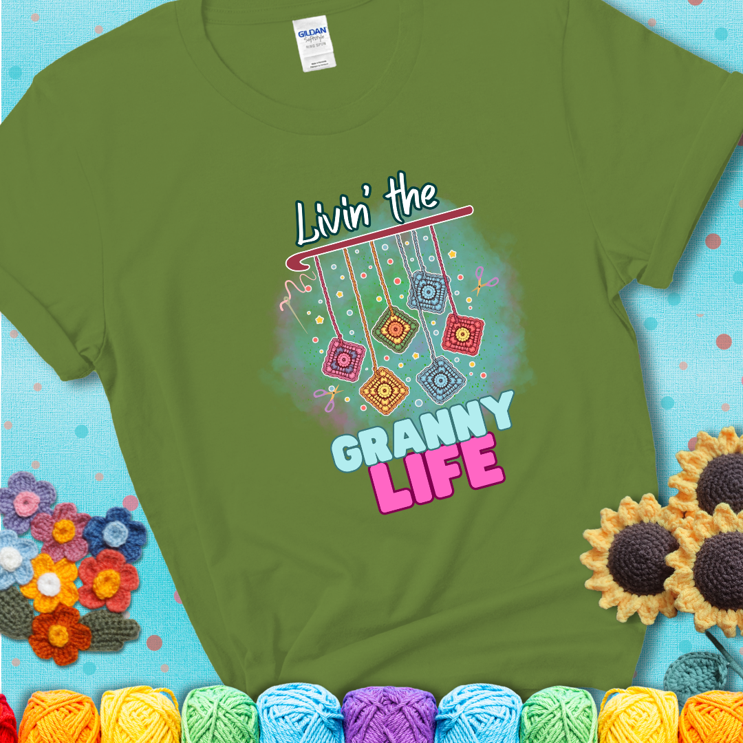 Green t-shirt shows colorful crochet granny squares hanging from a hook with text “Livin’ the Granny Life” in bold pink and blue font; a fun nod to classic crochet.