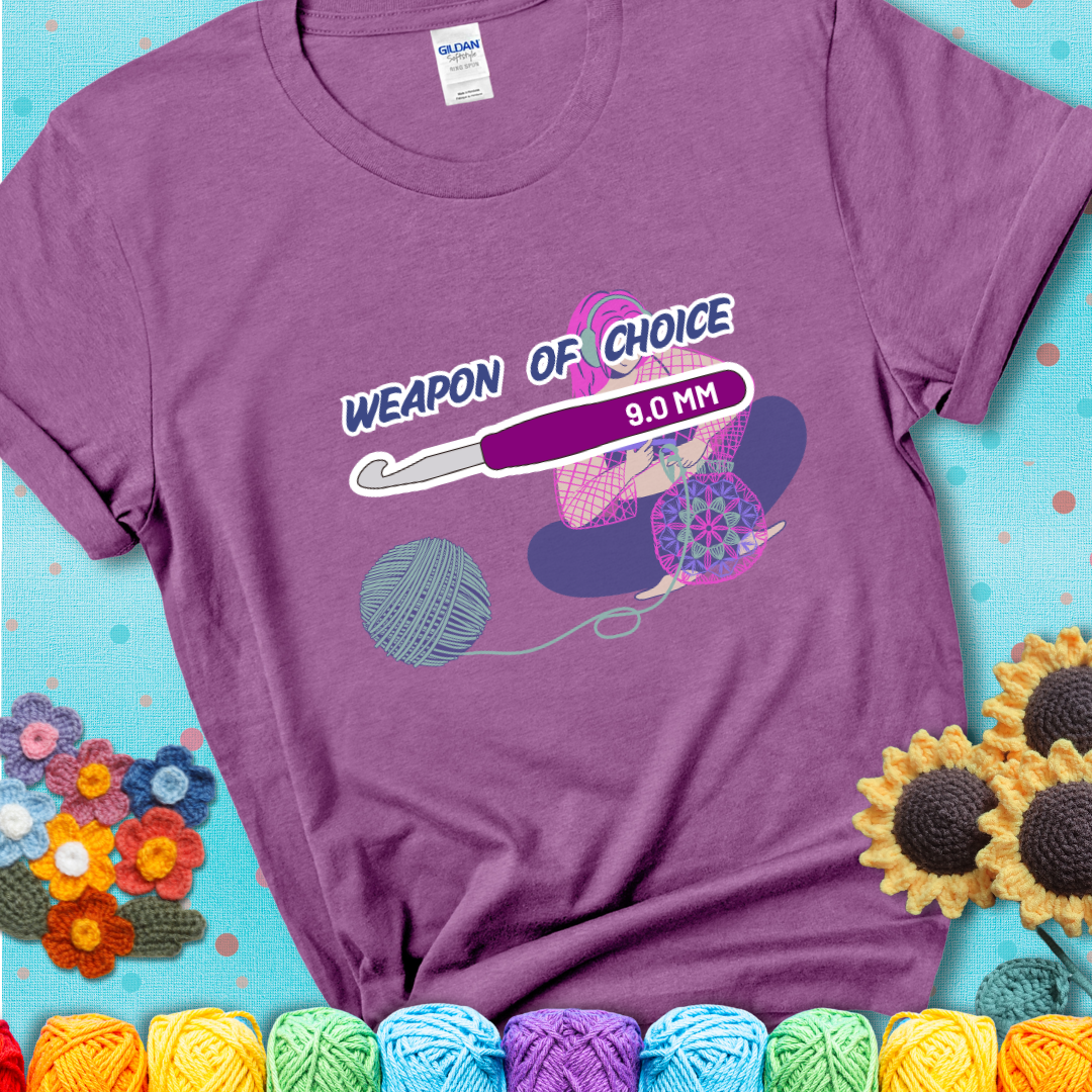 Light purple t-shirt with an Illustration of a crocheter sitting cross-legged with a 9.0 mm crochet hook above her, labeled “Weapon of Choice.” She’s crocheting a colorful mandala-style project from a large yarn ball.