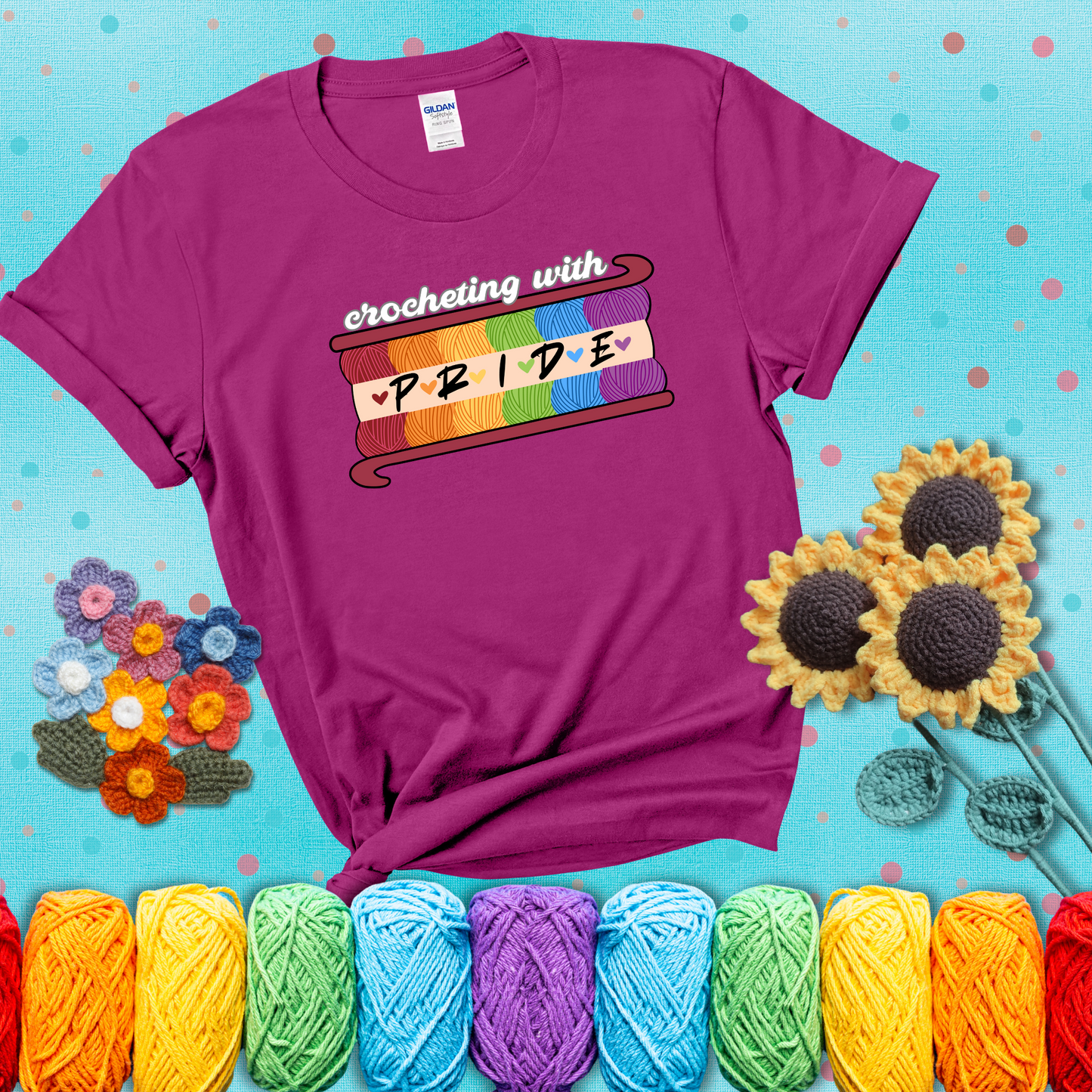 Yarn skeins in rainbow colors stacked in a hook shape. Text reads “Crocheting With Pride” showing a magenta tee with a teal sunflower background