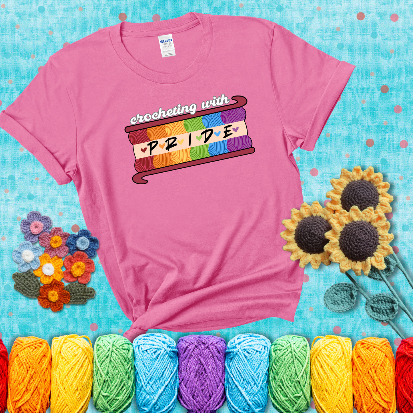 Yarn skeins in rainbow colors stacked in a hook shape. Text reads “Crocheting With Pride” showing a pink tee with a teal sunflower background