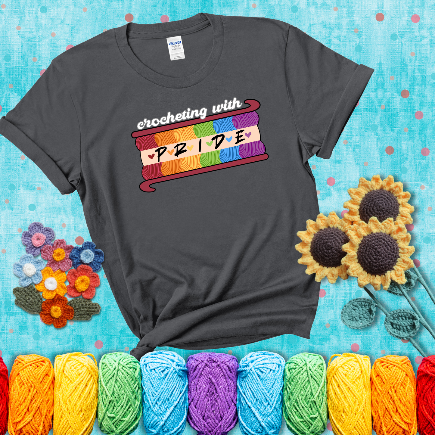 Yarn skeins in rainbow colors stacked in a hook shape. Text reads “Crocheting With Pride” showing a dark grey tee with a teal sunflower background