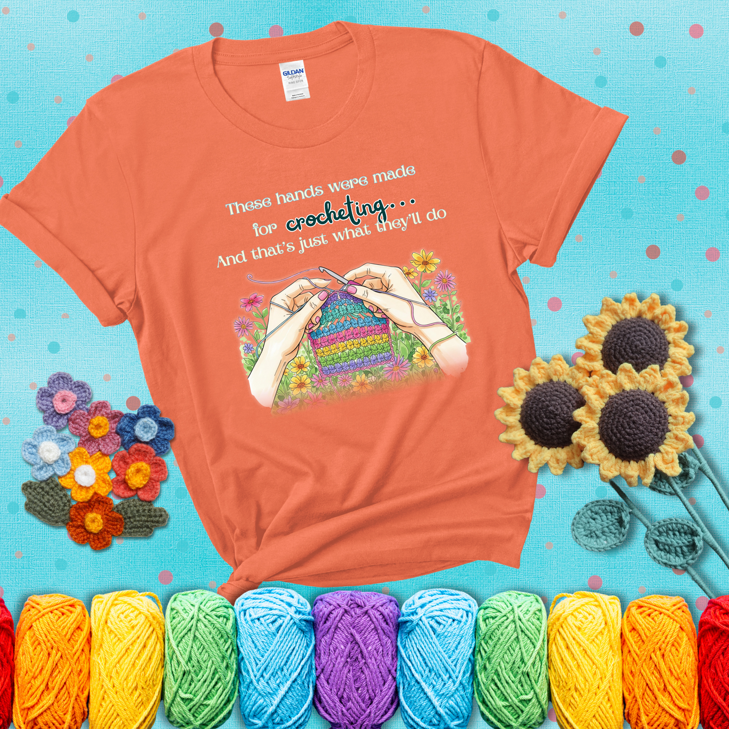 Illustration of two hands crocheting a rainbow swatch, surrounded by vibrant flowers with the phrase “These hands were made for crocheting… and that’s just what they’ll do,” showing a light orange tee with a teal sunflower background