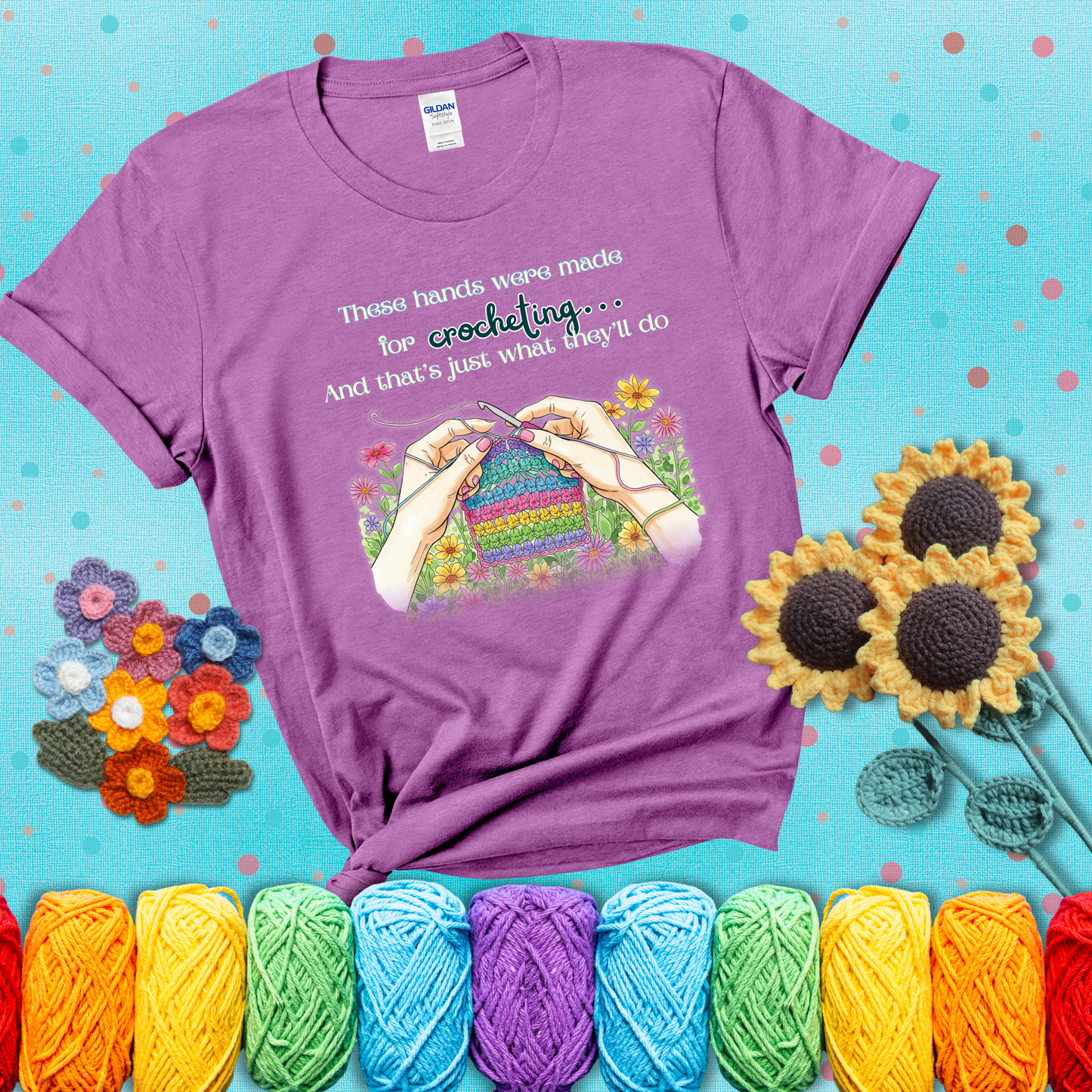 Illustration of two hands crocheting a rainbow swatch, surrounded by vibrant flowers with the phrase “These hands were made for crocheting… and that’s just what they’ll do,” showing a light purple tee with a teal sunflower background