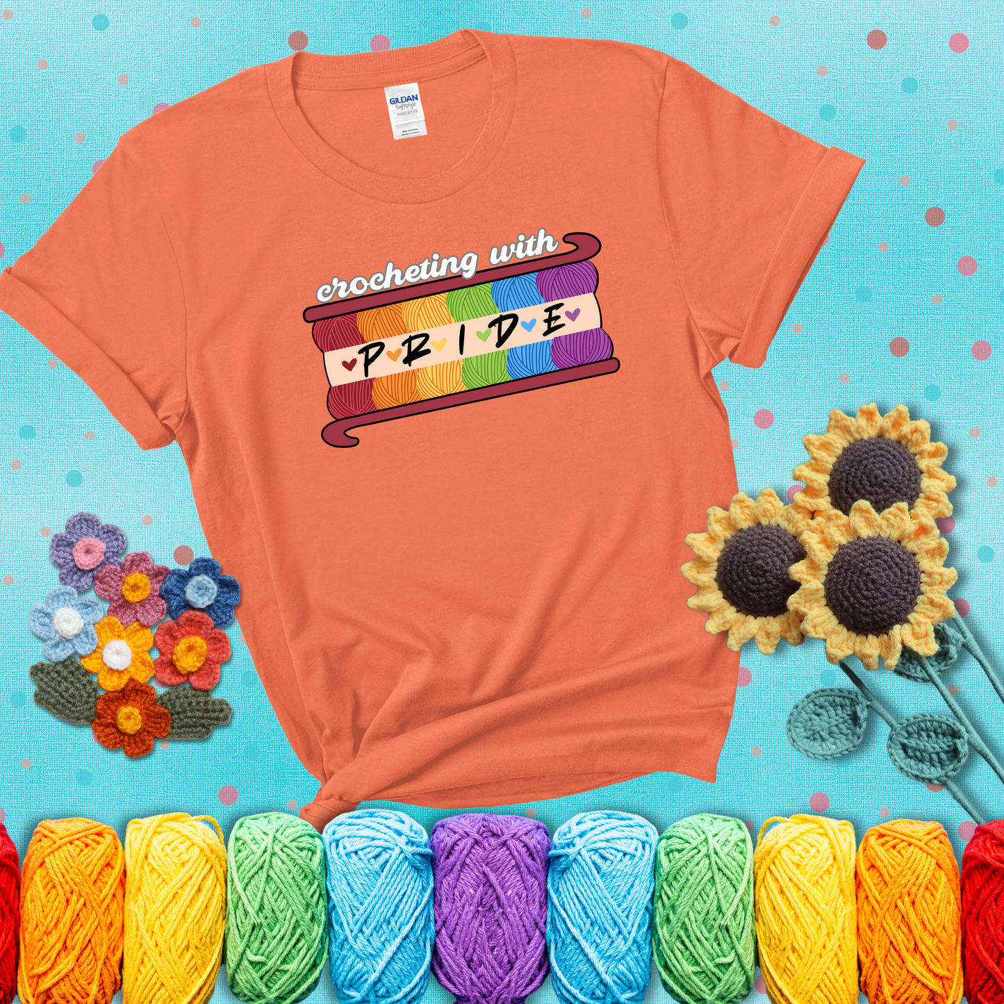 Yarn skeins in rainbow colors stacked in a hook shape. Text reads “Crocheting With Pride” showing a light orange tee with a teal sunflower background