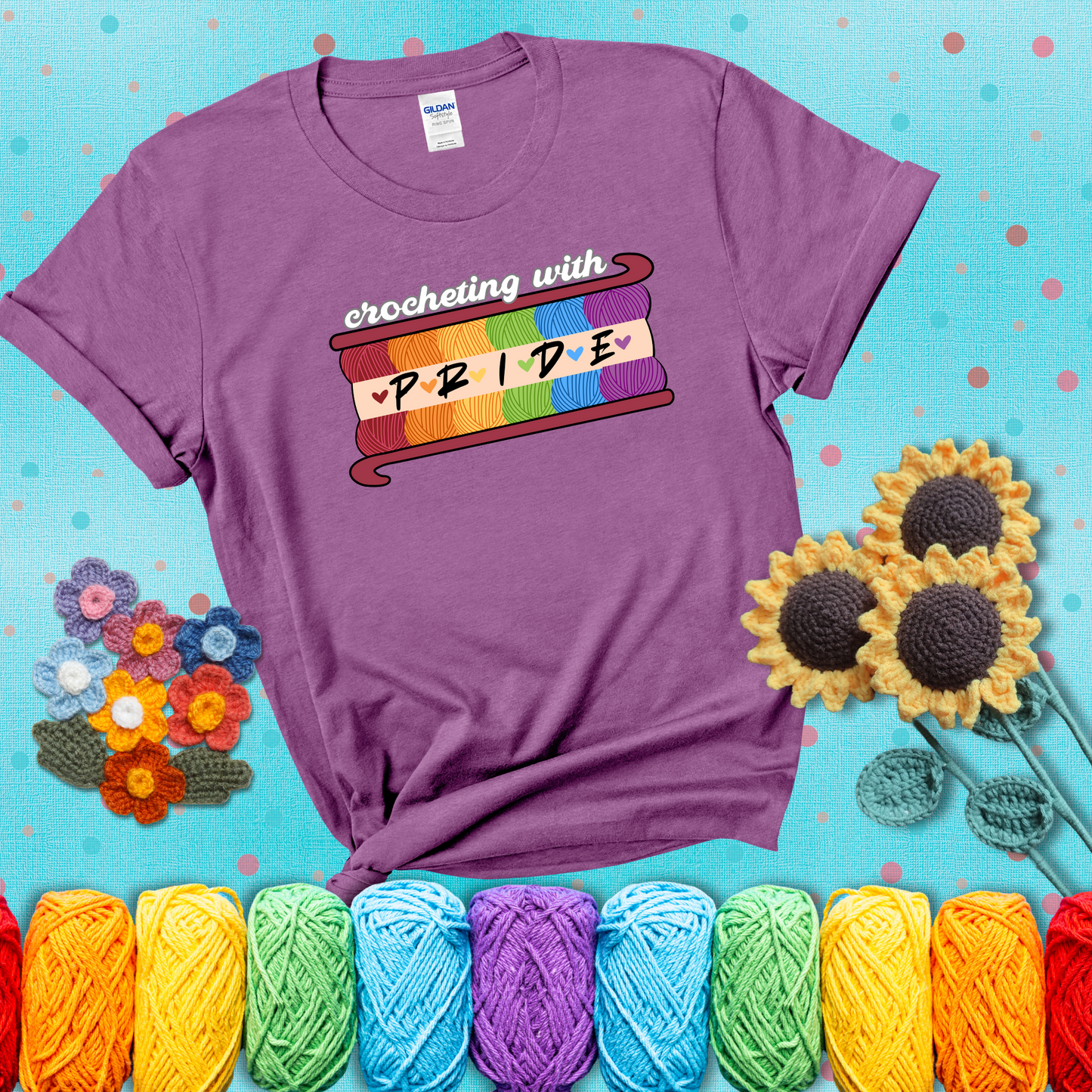 Yarn skeins in rainbow colors stacked in a hook shape. Text reads “Crocheting With Pride” showing a light purple tee with a teal sunflower background