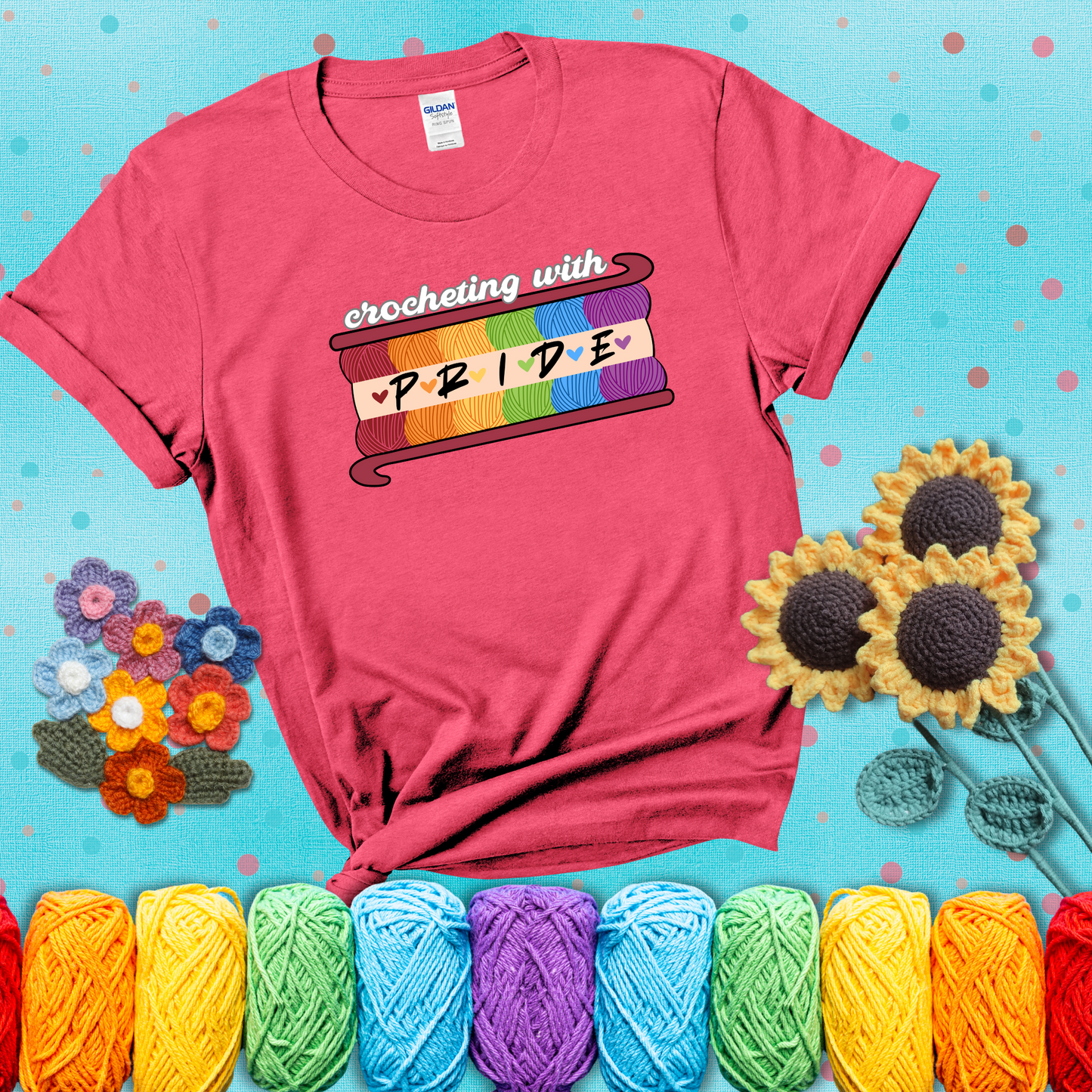 Yarn skeins in rainbow colors stacked in a hook shape. Text reads “Crocheting With Pride” showing a light red tee with a teal sunflower background