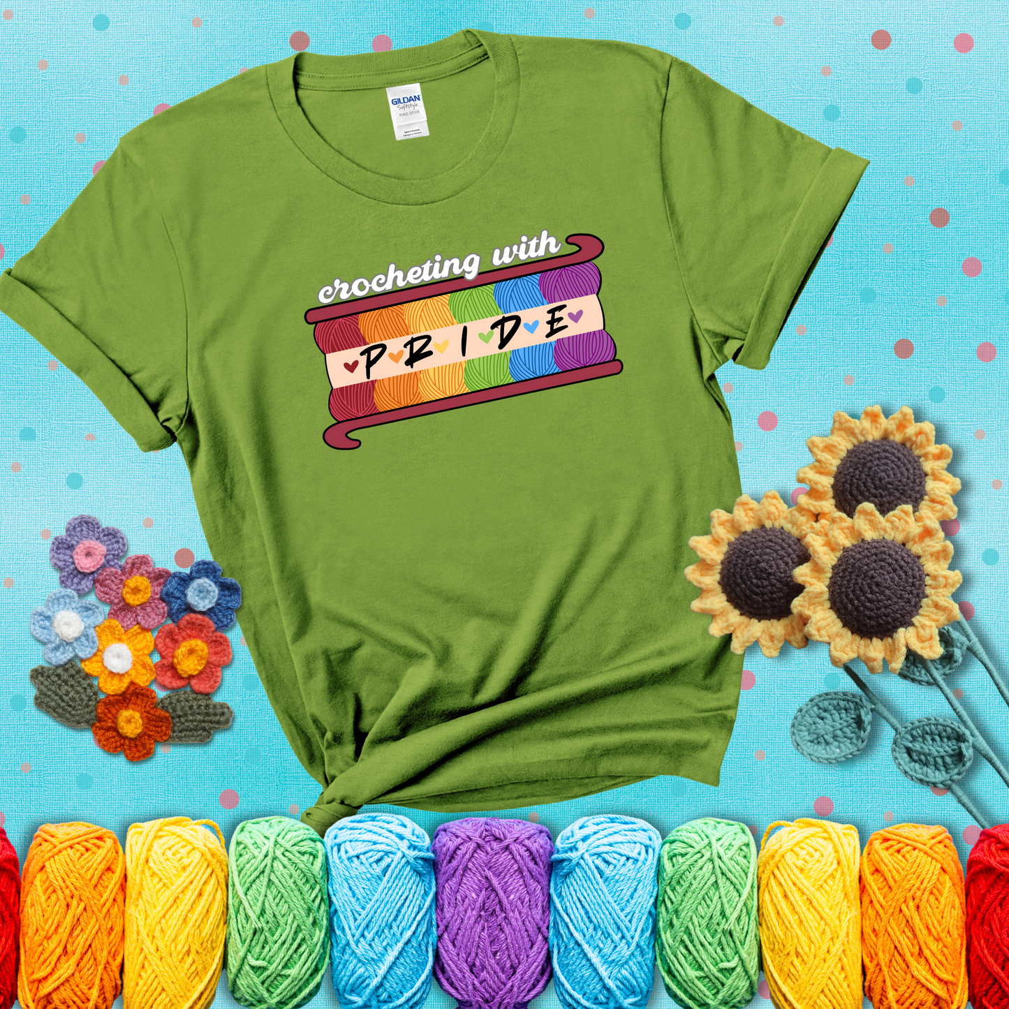 Yarn skeins in rainbow colors stacked in a hook shape. Text reads “Crocheting With Pride” showing a bright green tee with a teal sunflower background