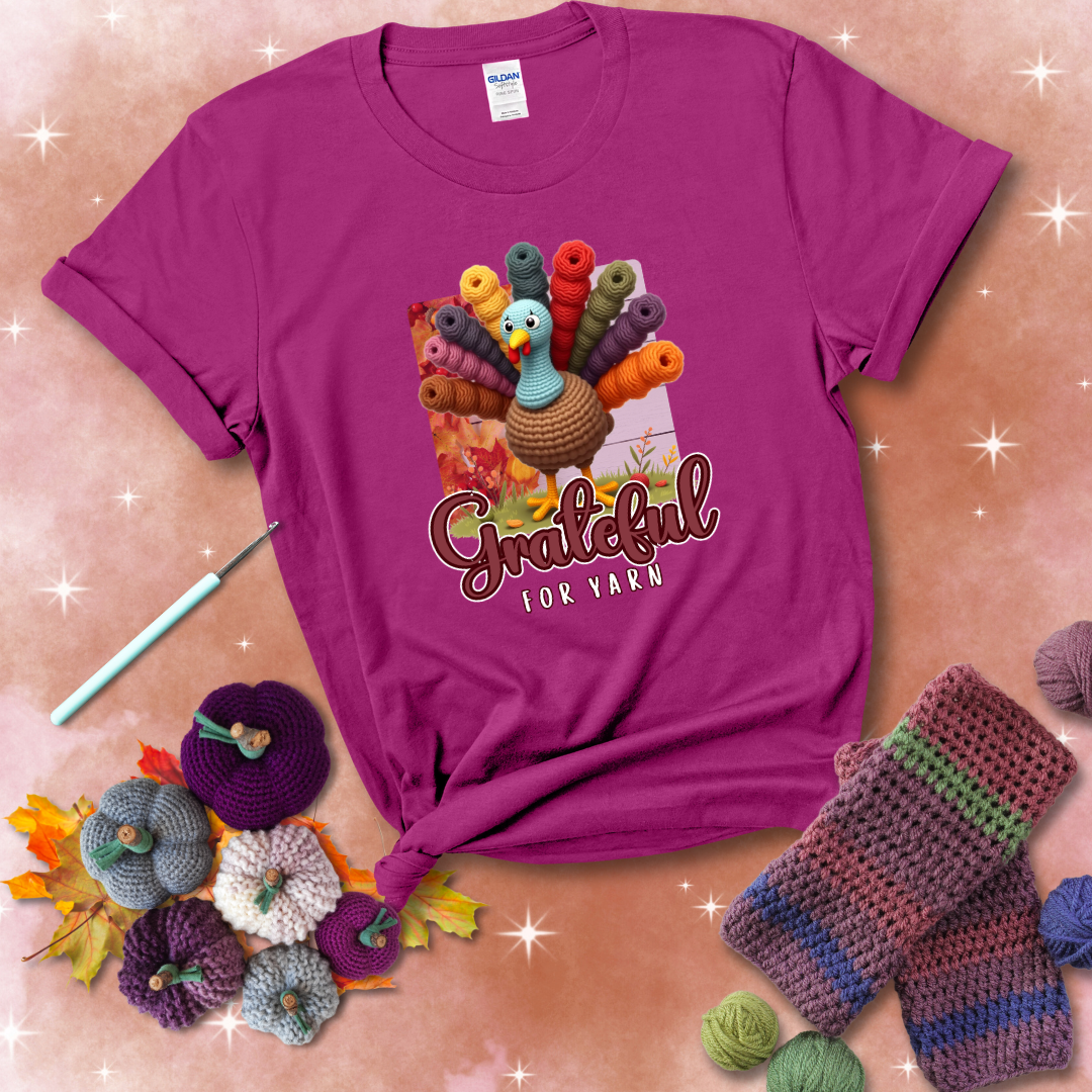 A crocheted turkey with colorful yarn skeins for tail feathers, standing on fall leaves with the text “Grateful for Yarn” showing a magenta tee with a festive Fall background