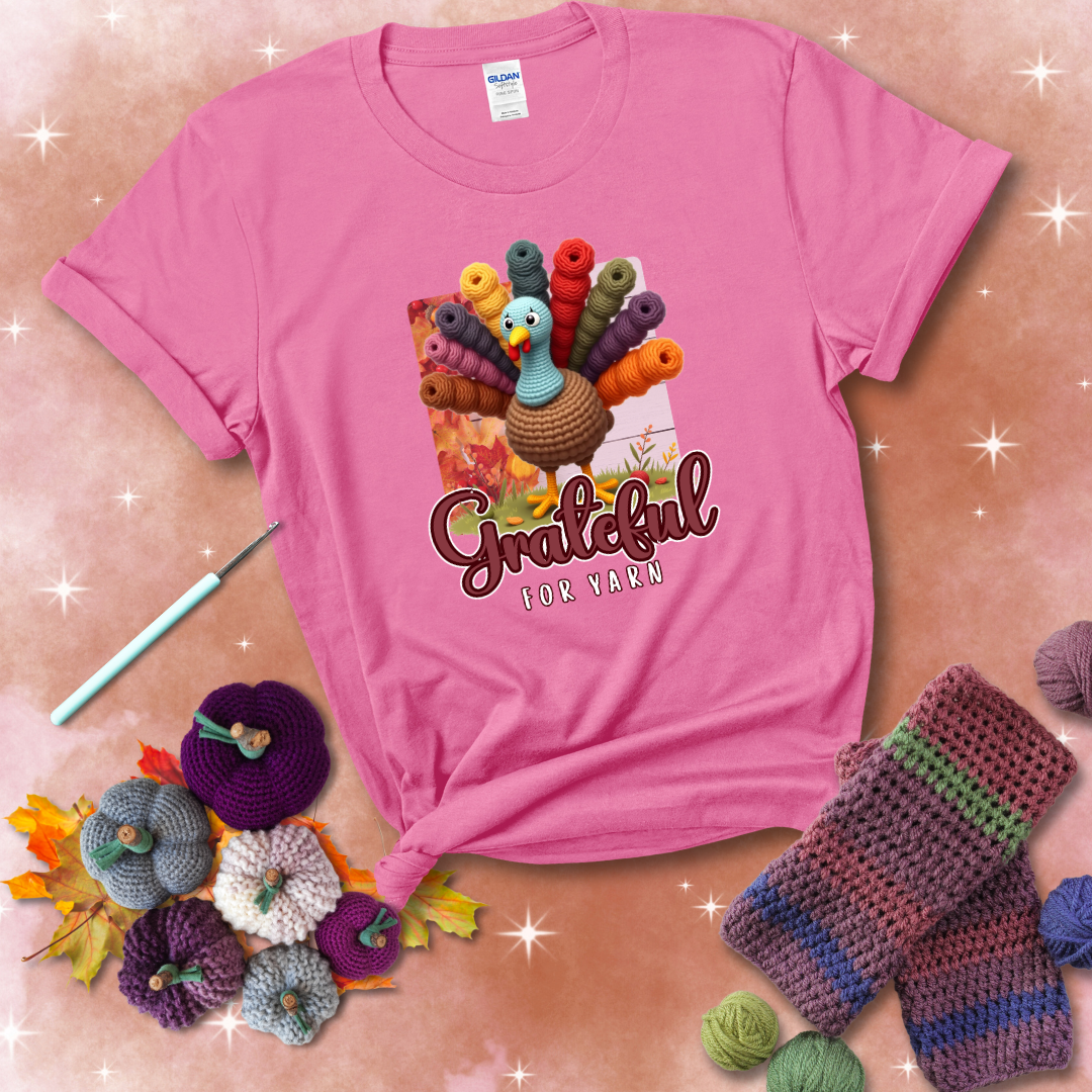 A crocheted turkey with colorful yarn skeins for tail feathers, standing on fall leaves with the text “Grateful for Yarn” showing a pink tee with a festive Fall background