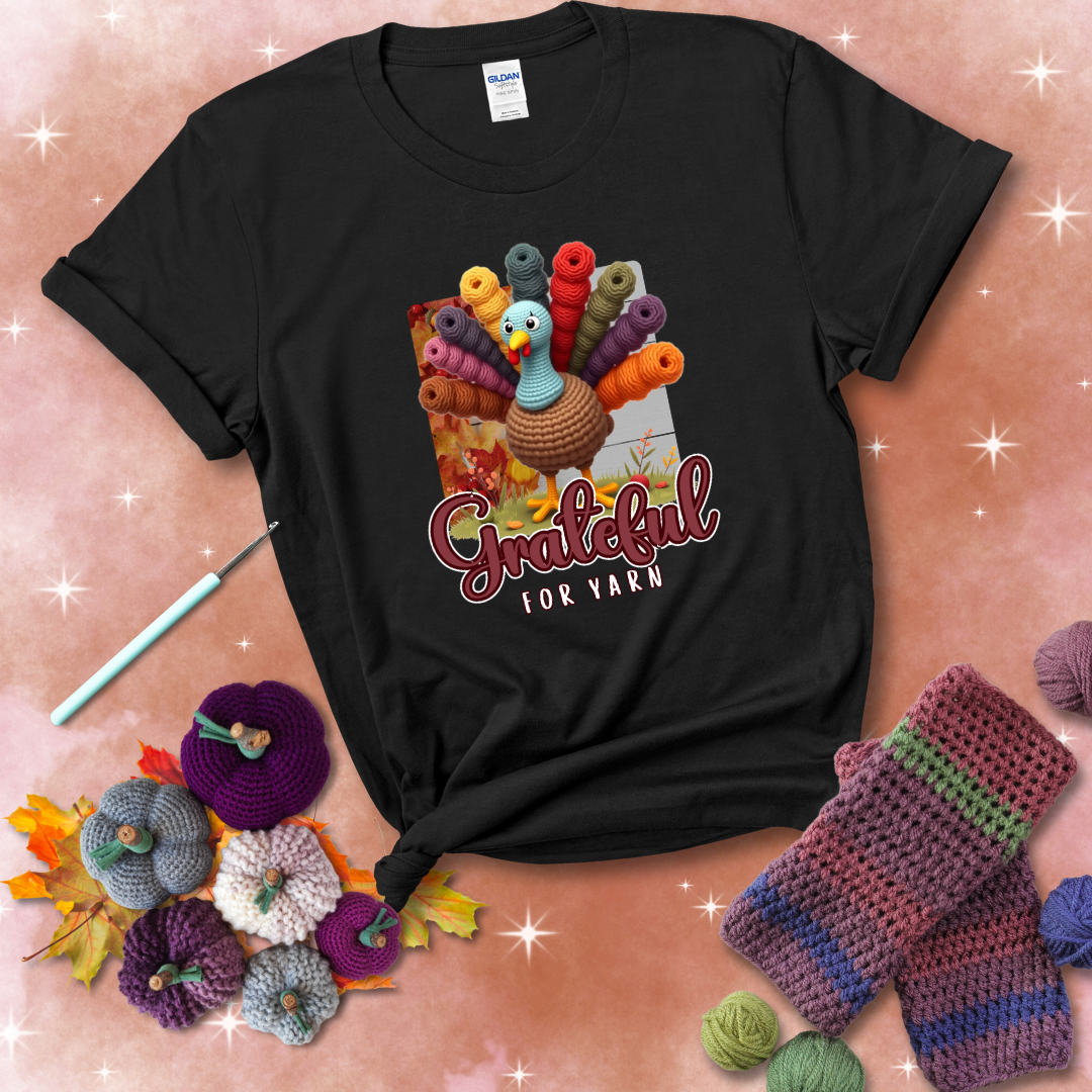A crocheted turkey with colorful yarn skeins for tail feathers, standing on fall leaves with the text “Grateful for Yarn” showing a black tee with a festive Fall background