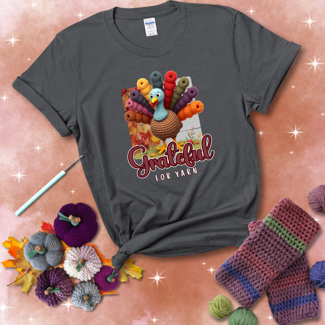 A crocheted turkey with colorful yarn skeins for tail feathers, standing on fall leaves with the text “Grateful for Yarn” showing a dark grey tee with a festive Fall background