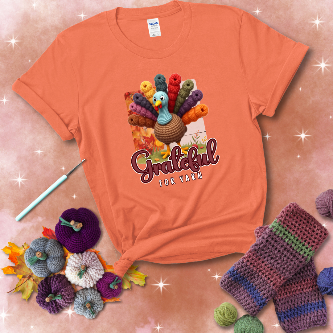 A crocheted turkey with colorful yarn skeins for tail feathers, standing on fall leaves with the text “Grateful for Yarn” showing a light orange tee with a festive Fall background