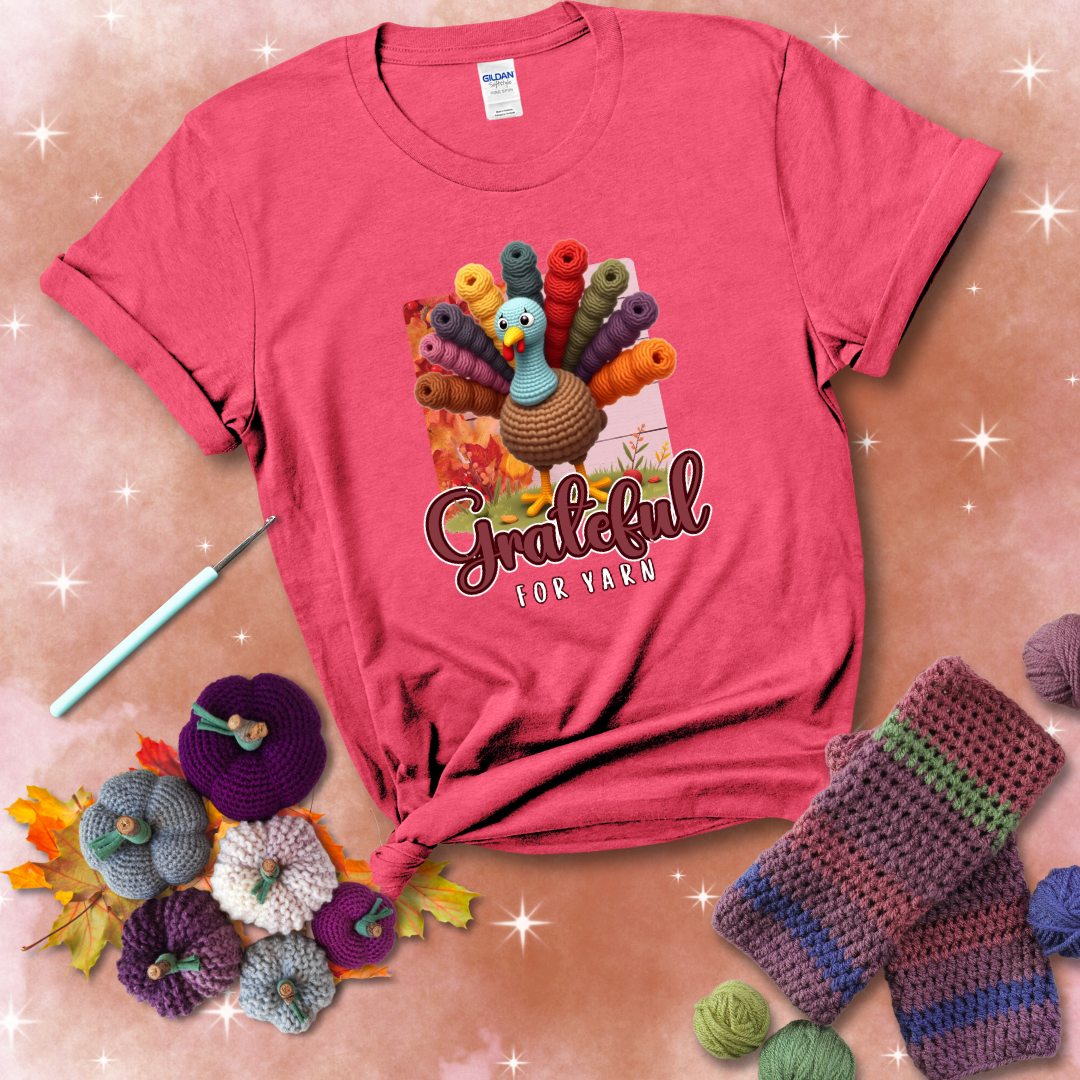 A crocheted turkey with colorful yarn skeins for tail feathers, standing on fall leaves with the text “Grateful for Yarn” showing a light red tee with a festive Fall background