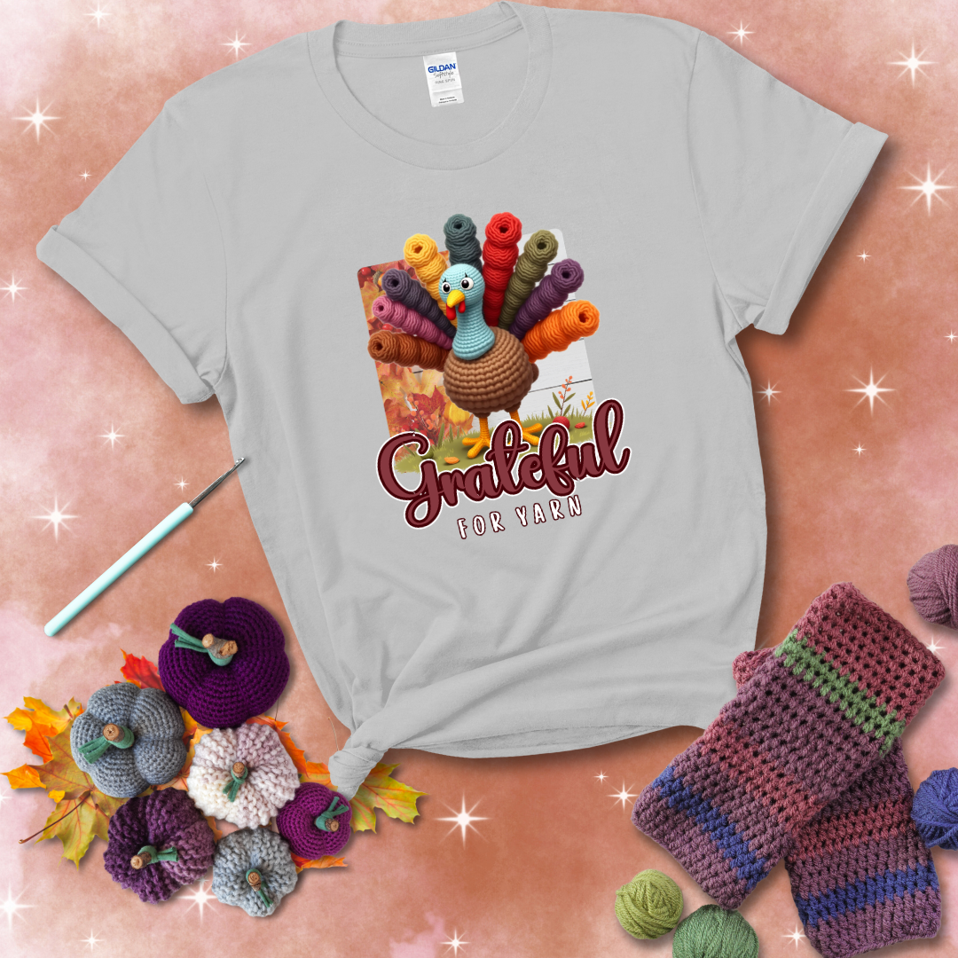 A crocheted turkey with colorful yarn skeins for tail feathers, standing on fall leaves with the text “Grateful for Yarn” showing a light grey tee with a festive Fall background