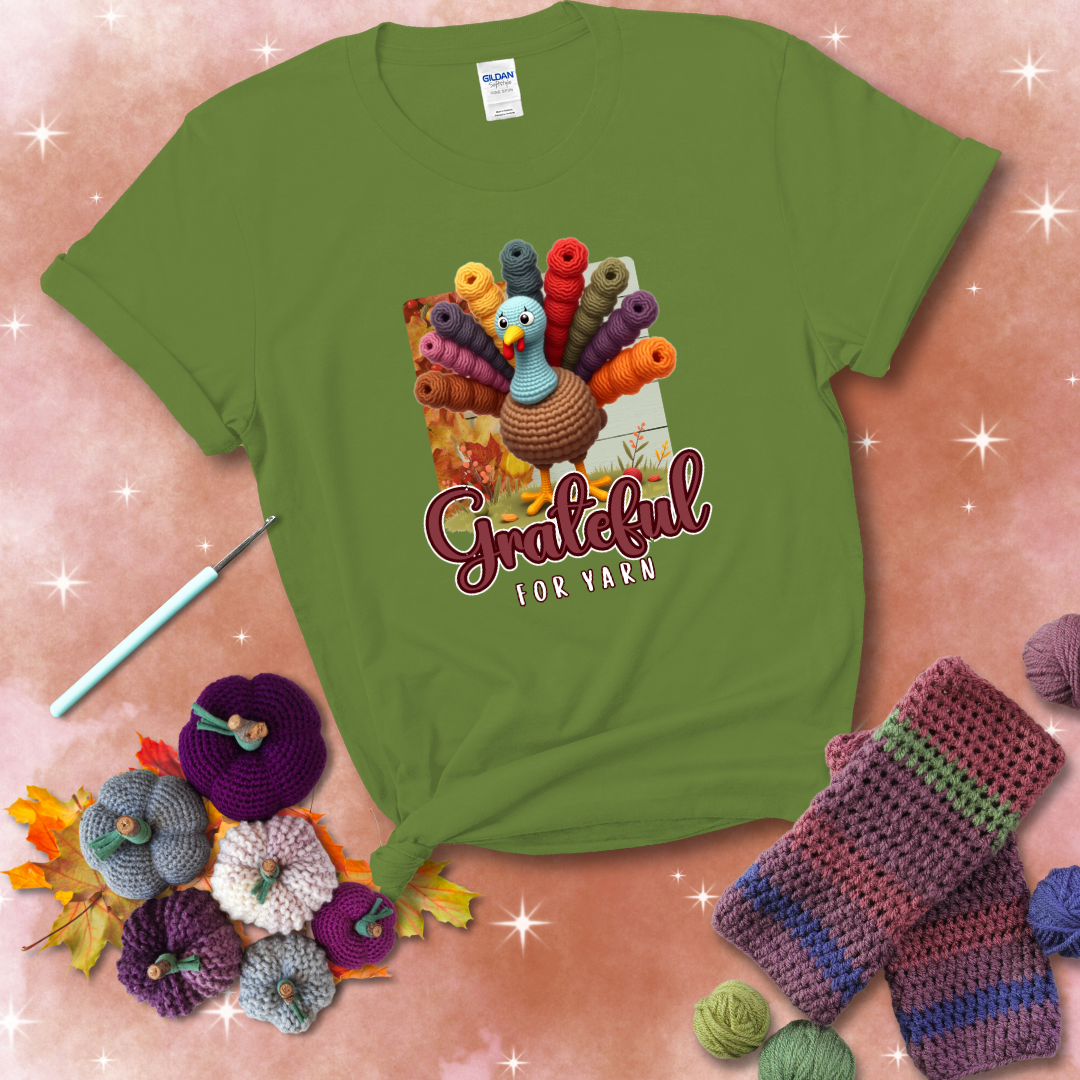 A crocheted turkey with colorful yarn skeins for tail feathers, standing on fall leaves with the text “Grateful for Yarn” showing a bright green tee with a festive Fall background