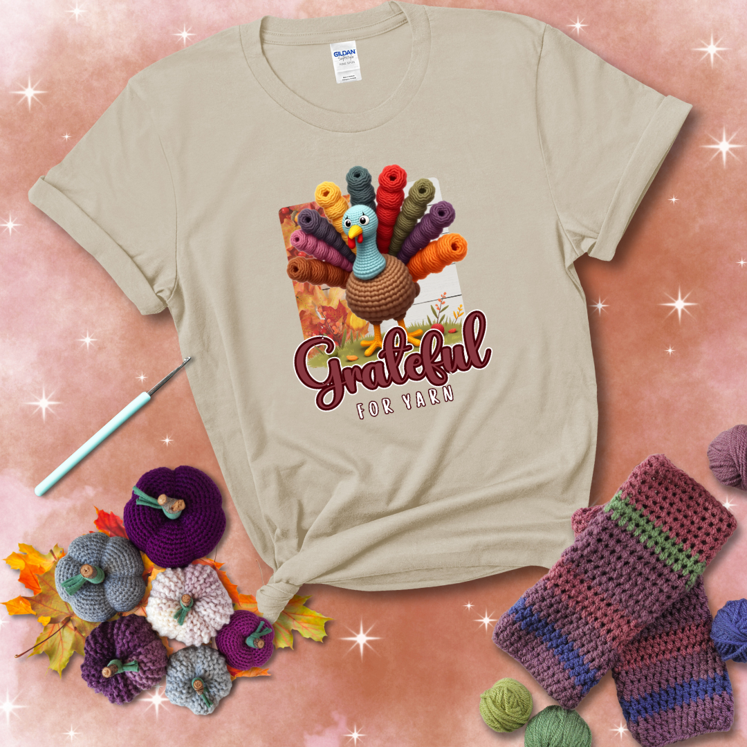 A crocheted turkey with colorful yarn skeins for tail feathers, standing on fall leaves with the text “Grateful for Yarn” showing a beige tee with a festive Fall background