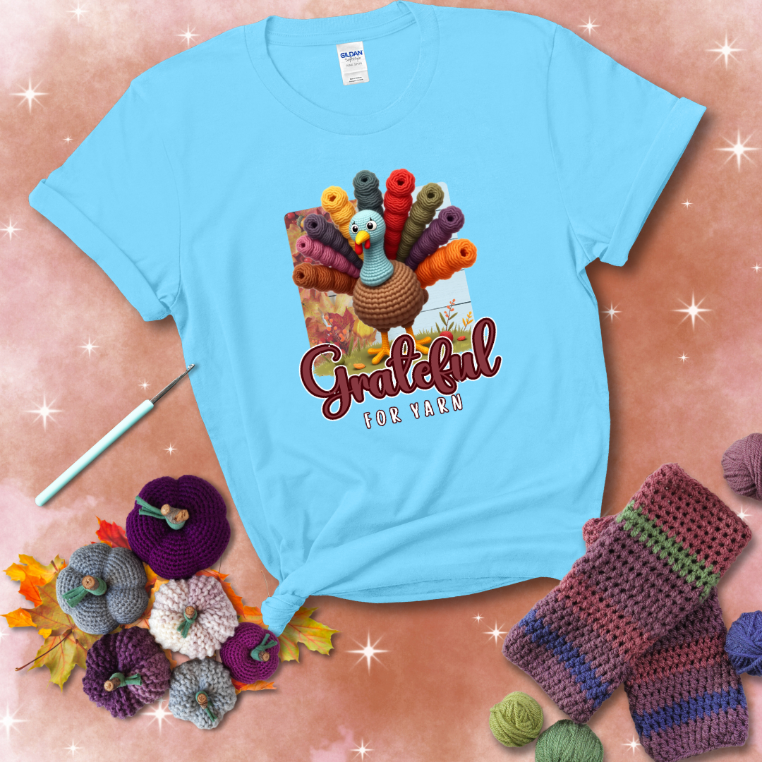 A crocheted turkey with colorful yarn skeins for tail feathers, standing on fall leaves with the text “Grateful for Yarn” showing a light blue tee with a festive Fall background