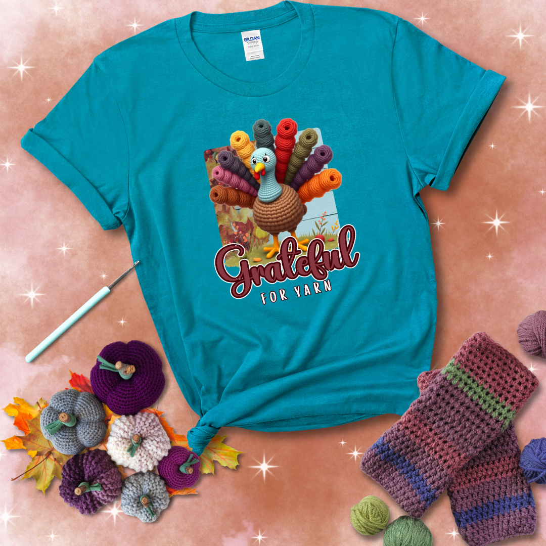 A crocheted turkey with colorful yarn skeins for tail feathers, standing on fall leaves with the text “Grateful for Yarn” showing a blue-green tee with a festive Fall background