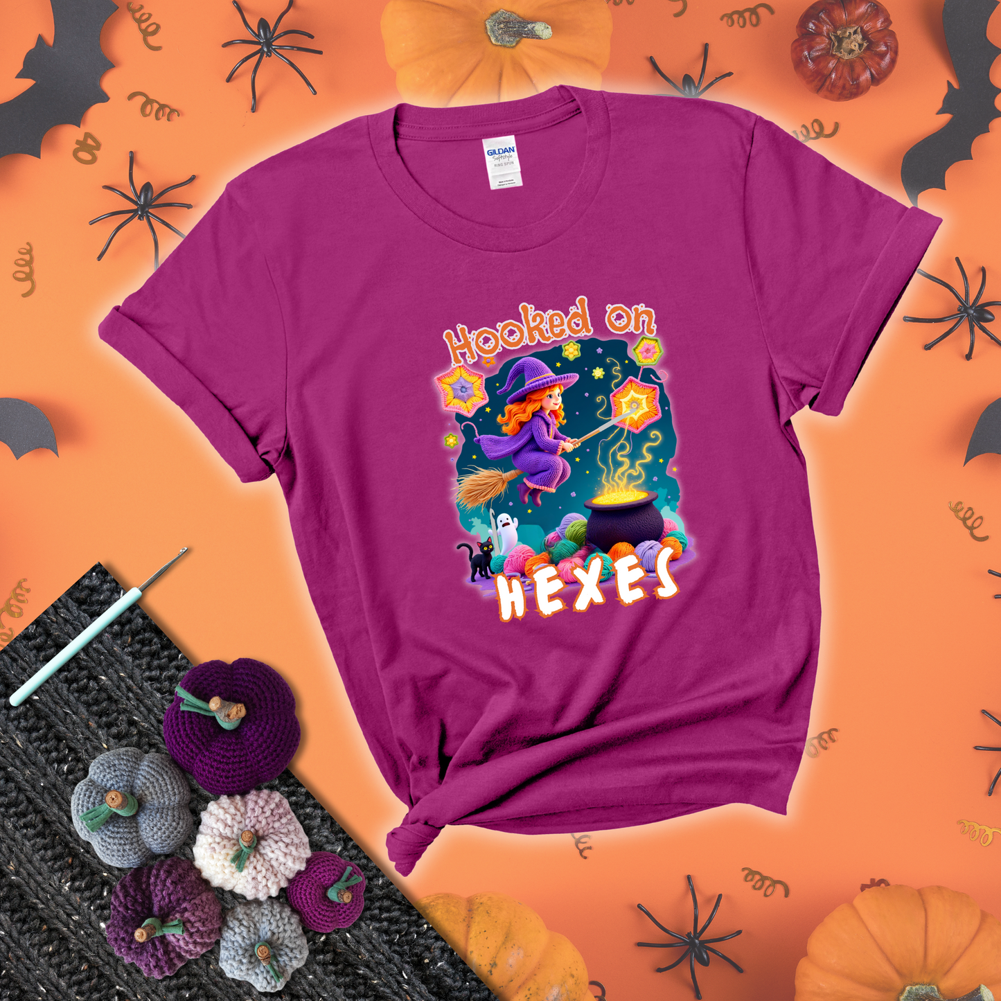 Witchy vibe with stars, hexagons, and a crochet hook. Text reads: “Hooked on Hexes.” A mystical and moody tee for fans of spooky stitches showing a magenta tee with a festive Halloween background