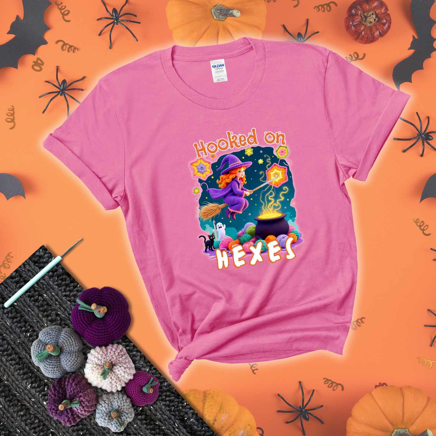 Witchy vibe with stars, hexagons, and a crochet hook. Text reads: “Hooked on Hexes.” A mystical and moody tee for fans of spooky stitches showing a pink tee with a festive Halloween background