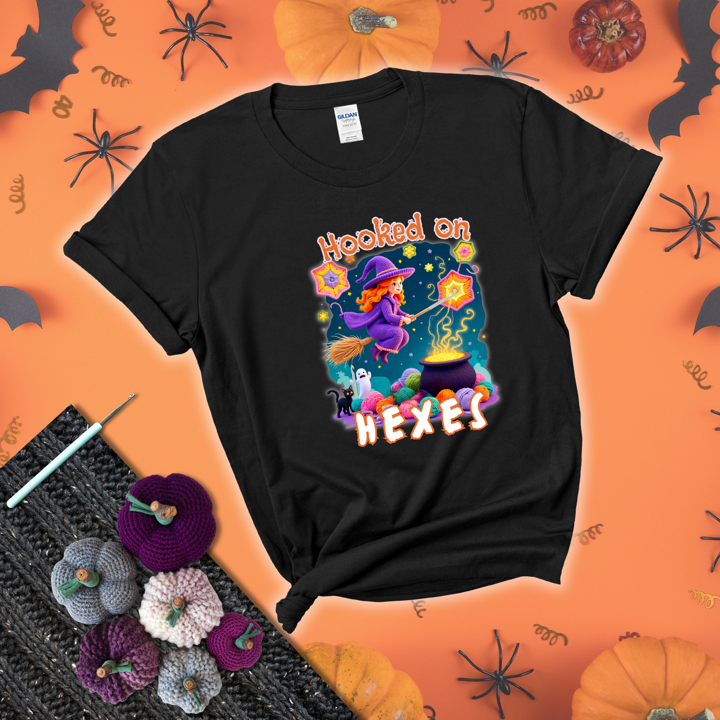 Witchy vibe with stars, hexagons, and a crochet hook. Text reads: “Hooked on Hexes.” A mystical and moody tee for fans of spooky stitches showing a black tee with a festive Halloween background