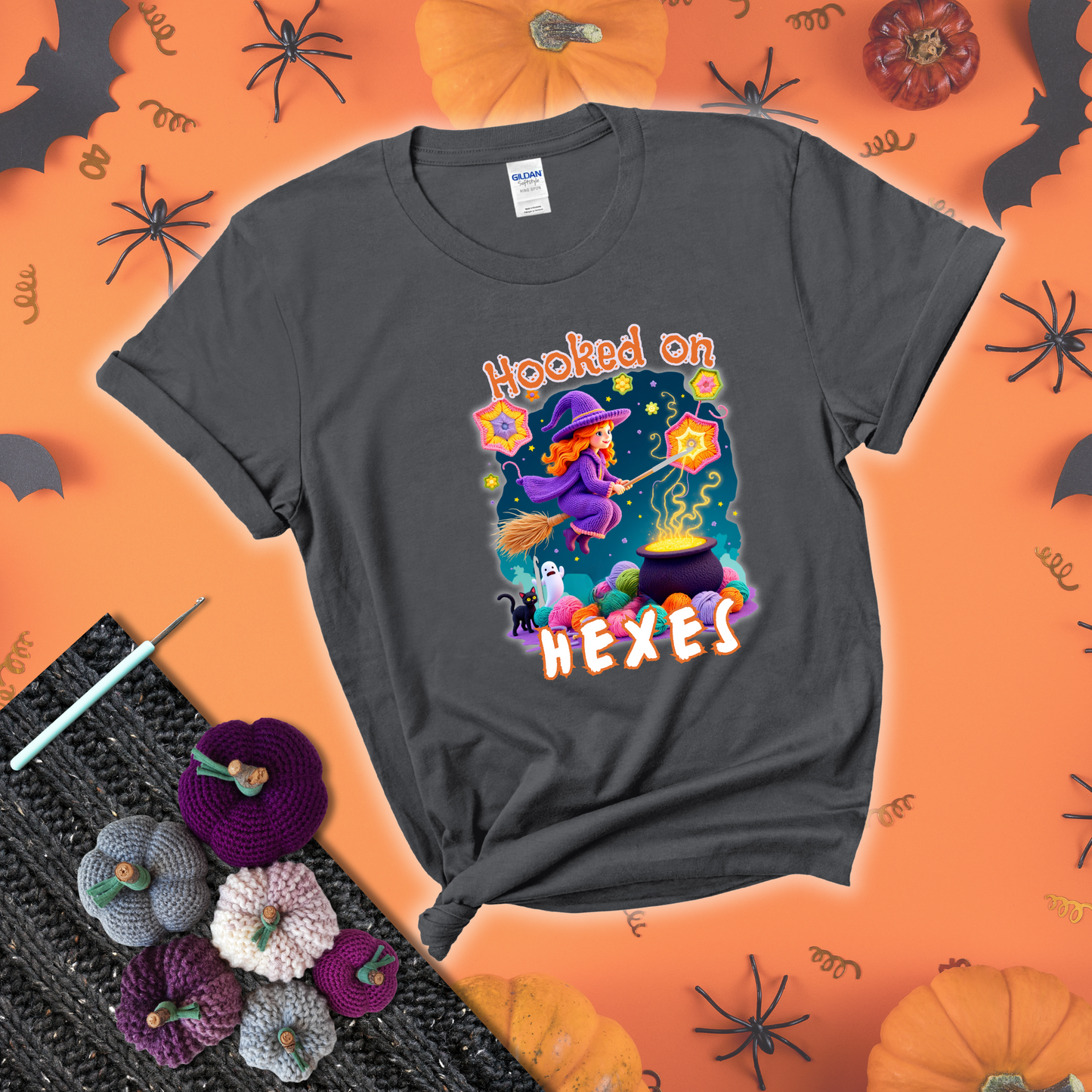 Witchy vibe with stars, hexagons, and a crochet hook. Text reads: “Hooked on Hexes.” A mystical and moody tee for fans of spooky stitches showing a dark grey tee with a festive Halloween background