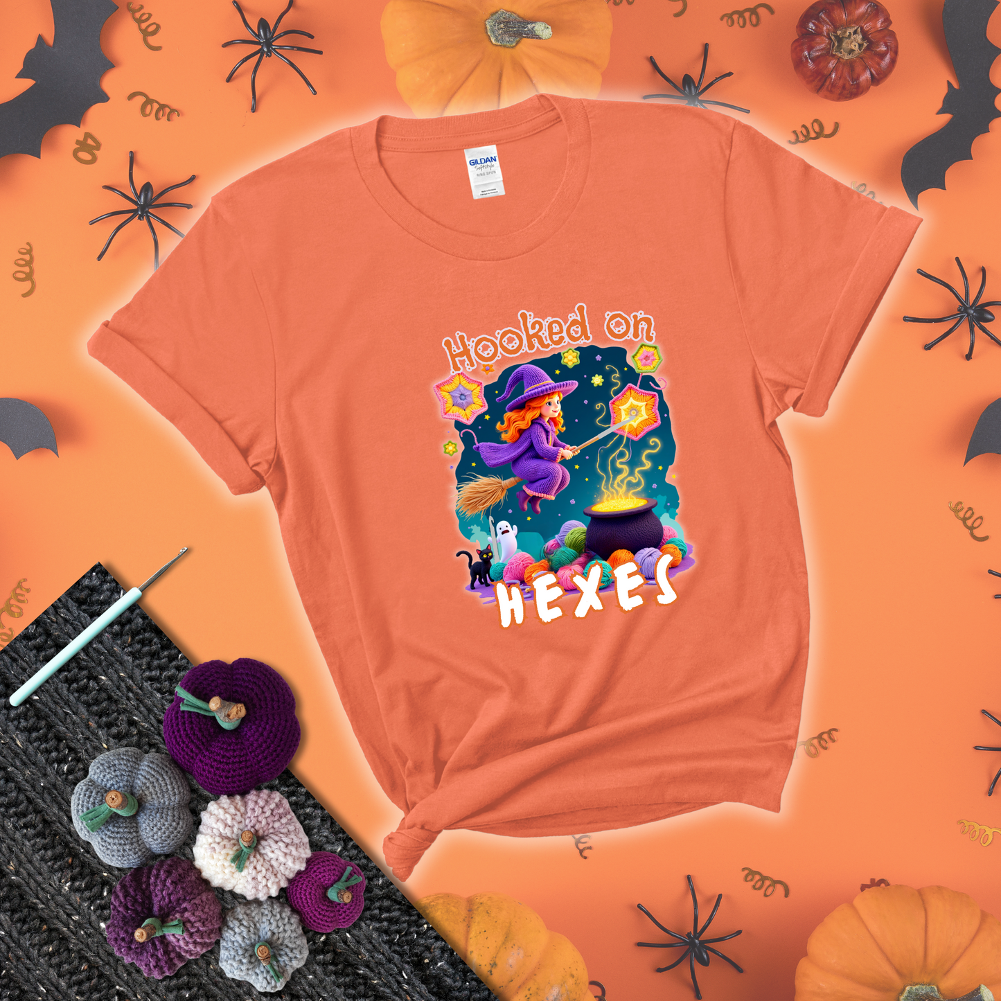 Witchy vibe with stars, hexagons, and a crochet hook. Text reads: “Hooked on Hexes.” A mystical and moody tee for fans of spooky stitches showing a light orange tee with a festive Halloween background