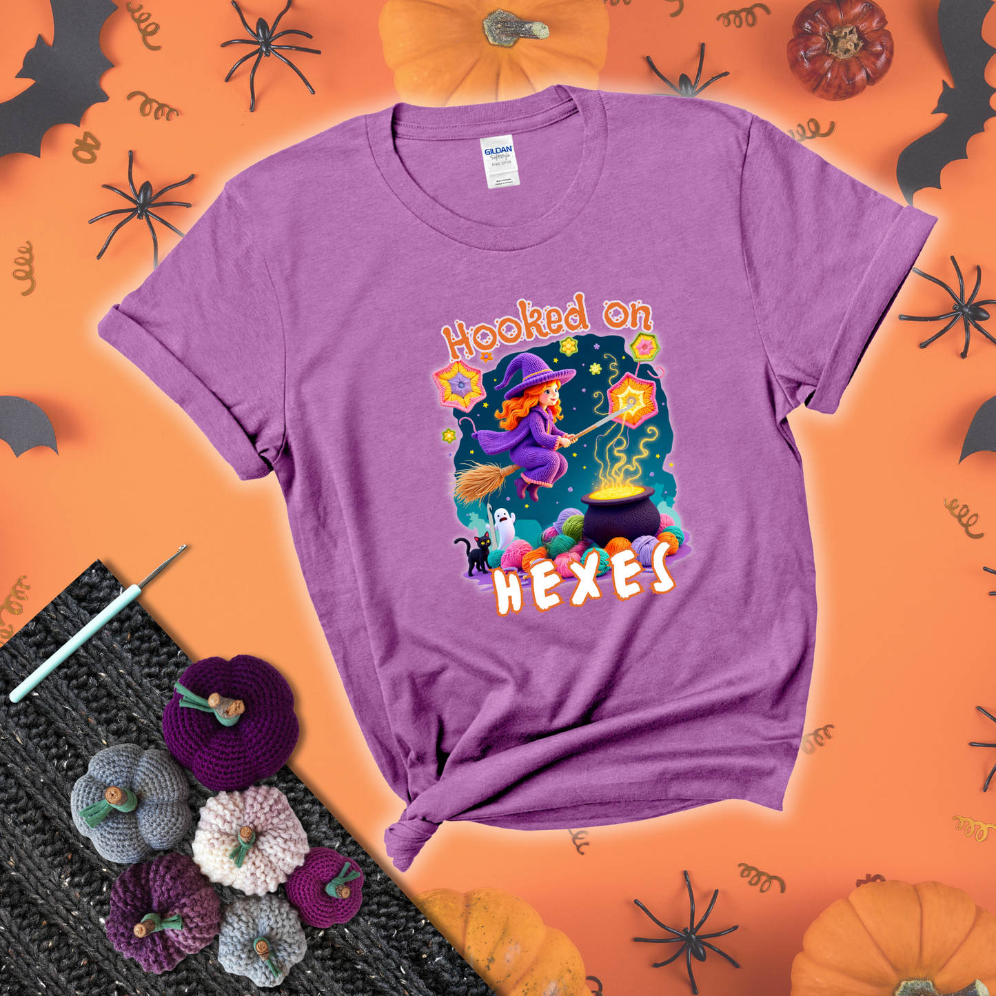 Witchy vibe with stars, hexagons, and a crochet hook. Text reads: “Hooked on Hexes.” A mystical and moody tee for fans of spooky stitches showing a light purple tee with a festive Halloween background