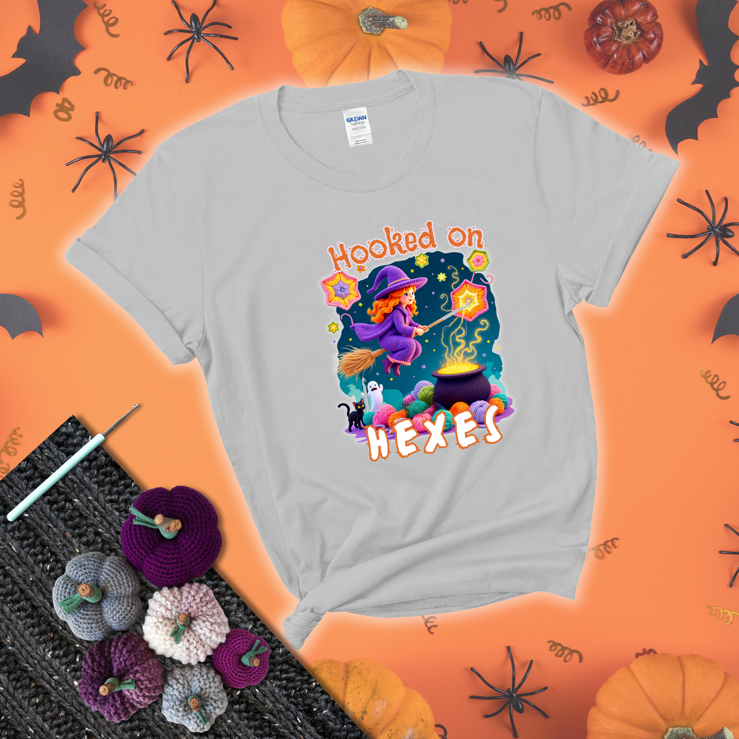 Witchy vibe with stars, hexagons, and a crochet hook. Text reads: “Hooked on Hexes.” A mystical and moody tee for fans of spooky stitches showing a light grey tee with a festive Halloween background