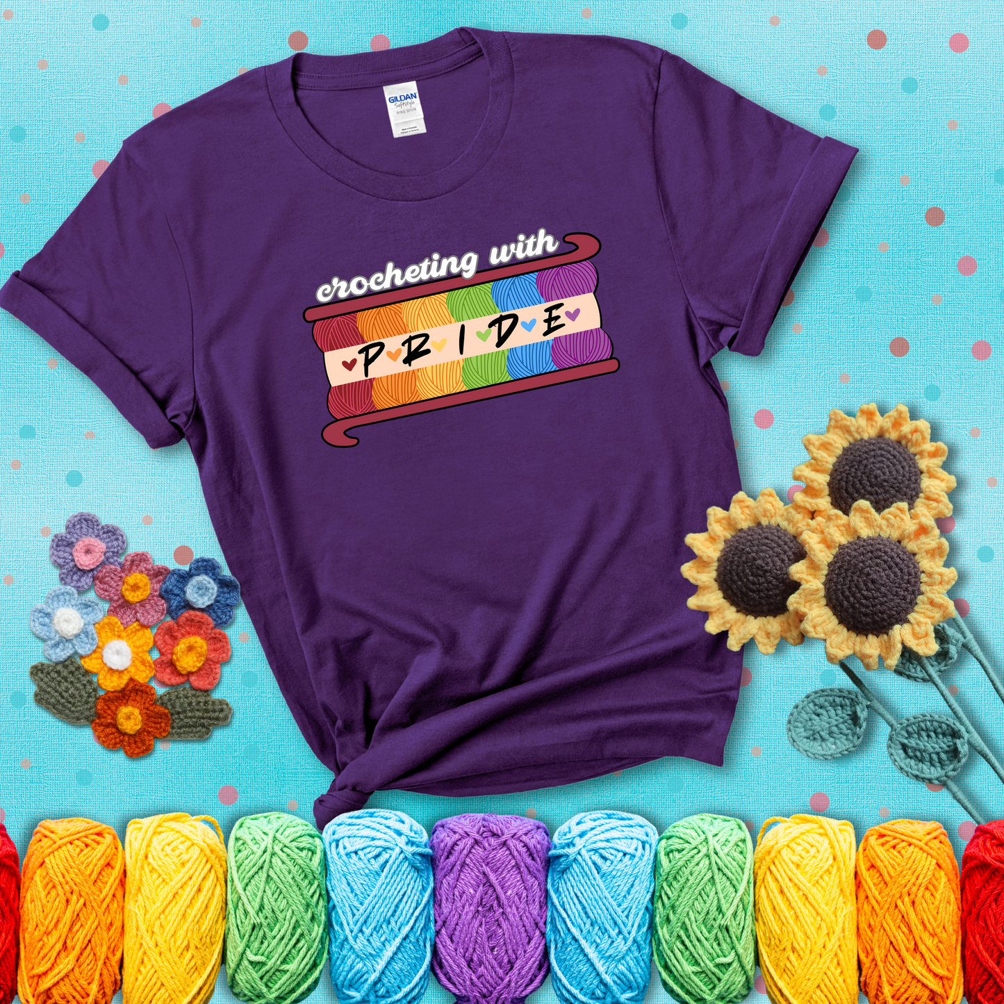 Yarn skeins in rainbow colors stacked in a hook shape. Text reads “Crocheting With Pride” showing a purple tee with a teal sunflower background