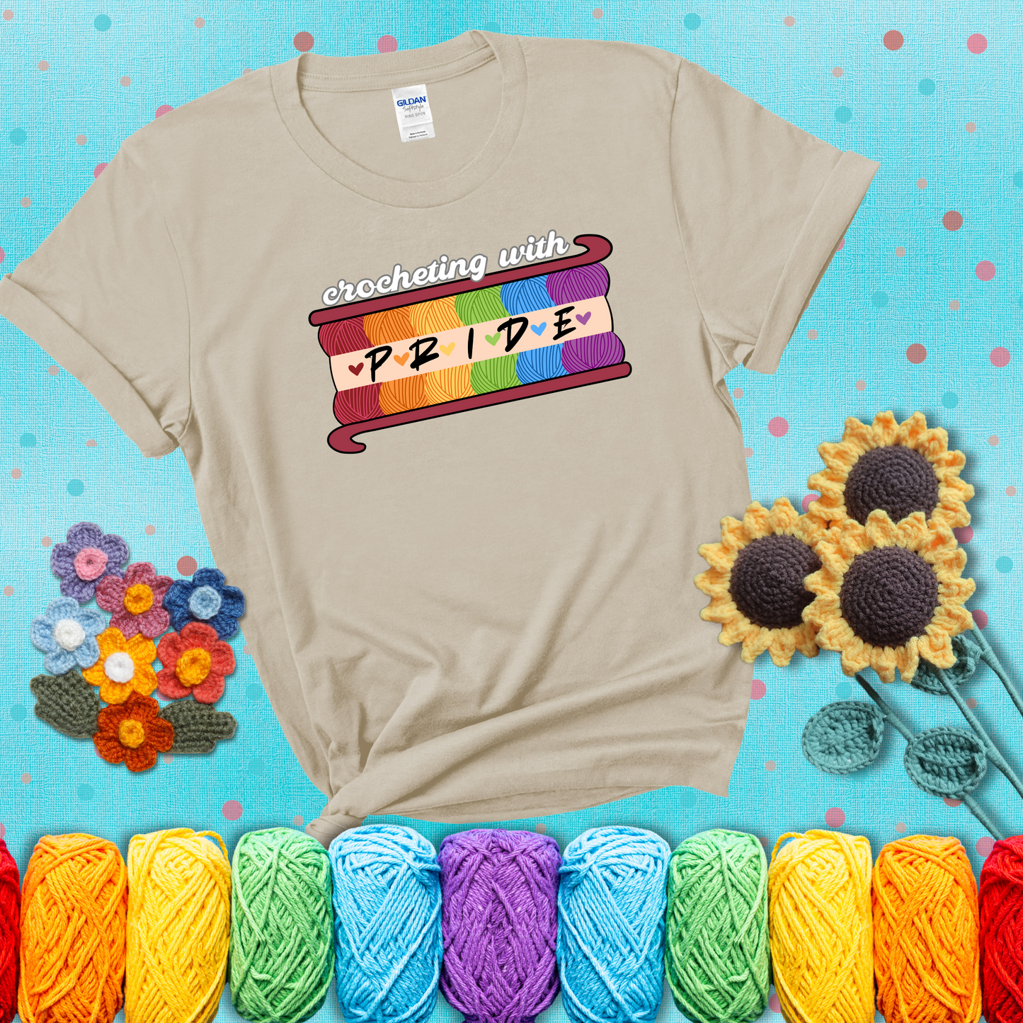 Yarn skeins in rainbow colors stacked in a hook shape. Text reads “Crocheting With Pride” showing a beige tee with a teal sunflower background