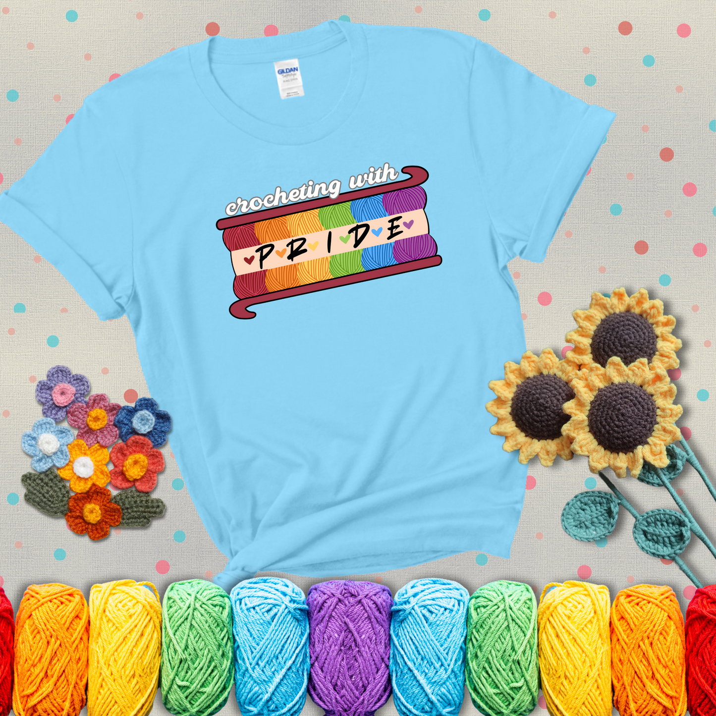 Yarn skeins in rainbow colors stacked in a hook shape. Text reads “Crocheting With Pride” showing a light blue tee with grey sunflower background