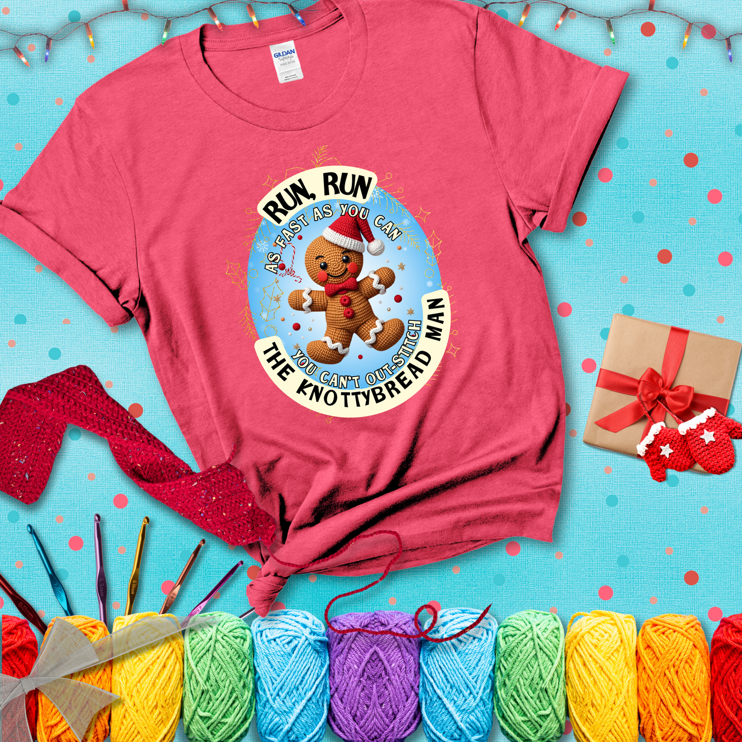 Cute crocheted gingerbread man in Santa hat with playful text: “Run, Run As Fast As You Can, You Can’t Out-Stitch the Knottybread Man.” A festive crochet pun shirt, showing a light red tee with a festive Christmas background
