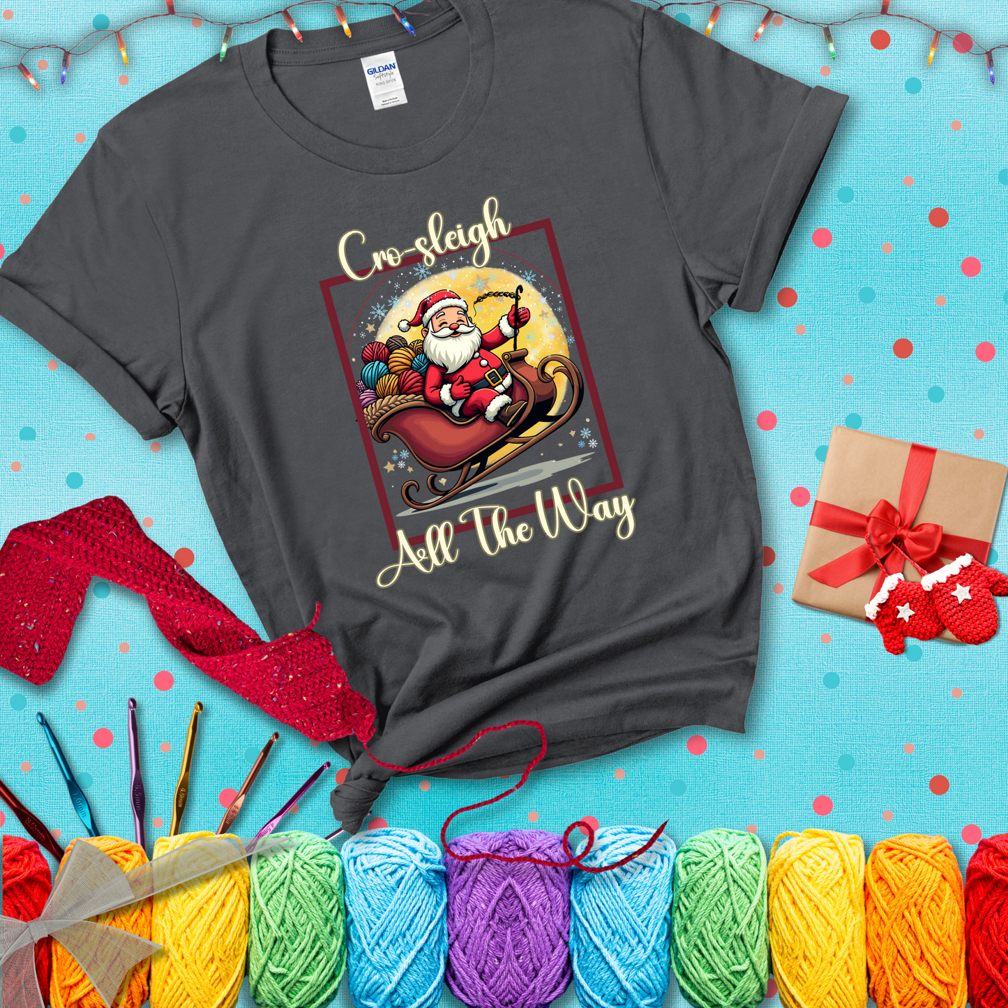 Jolly Santa rides a sleigh overflowing with colorful yarn balls, surrounded by sparkles and holiday cheer. Text reads “Cro-sleigh All the Way" showing a dark grey tee with a festive Christmas background