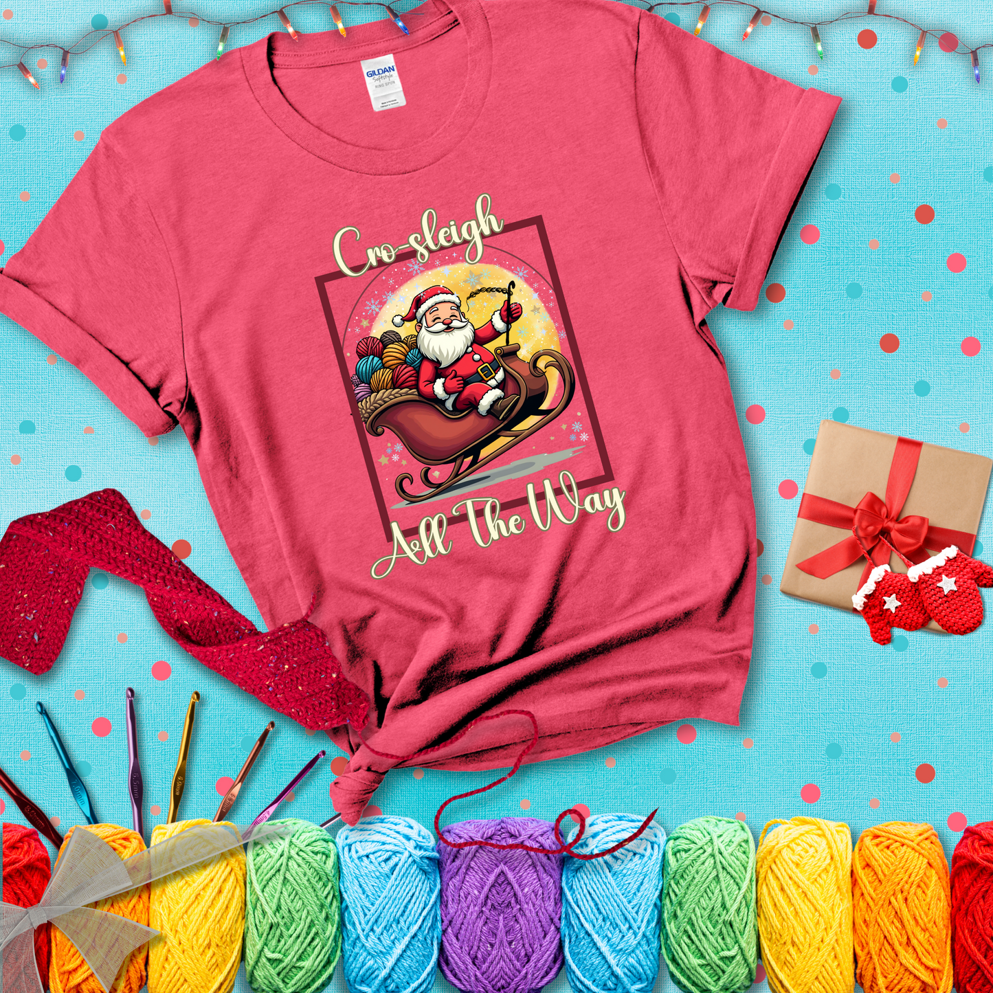 Jolly Santa rides a sleigh overflowing with colorful yarn balls, surrounded by sparkles and holiday cheer. Text reads “Cro-sleigh All the Way" showing a light red tee with a festive Christmas background