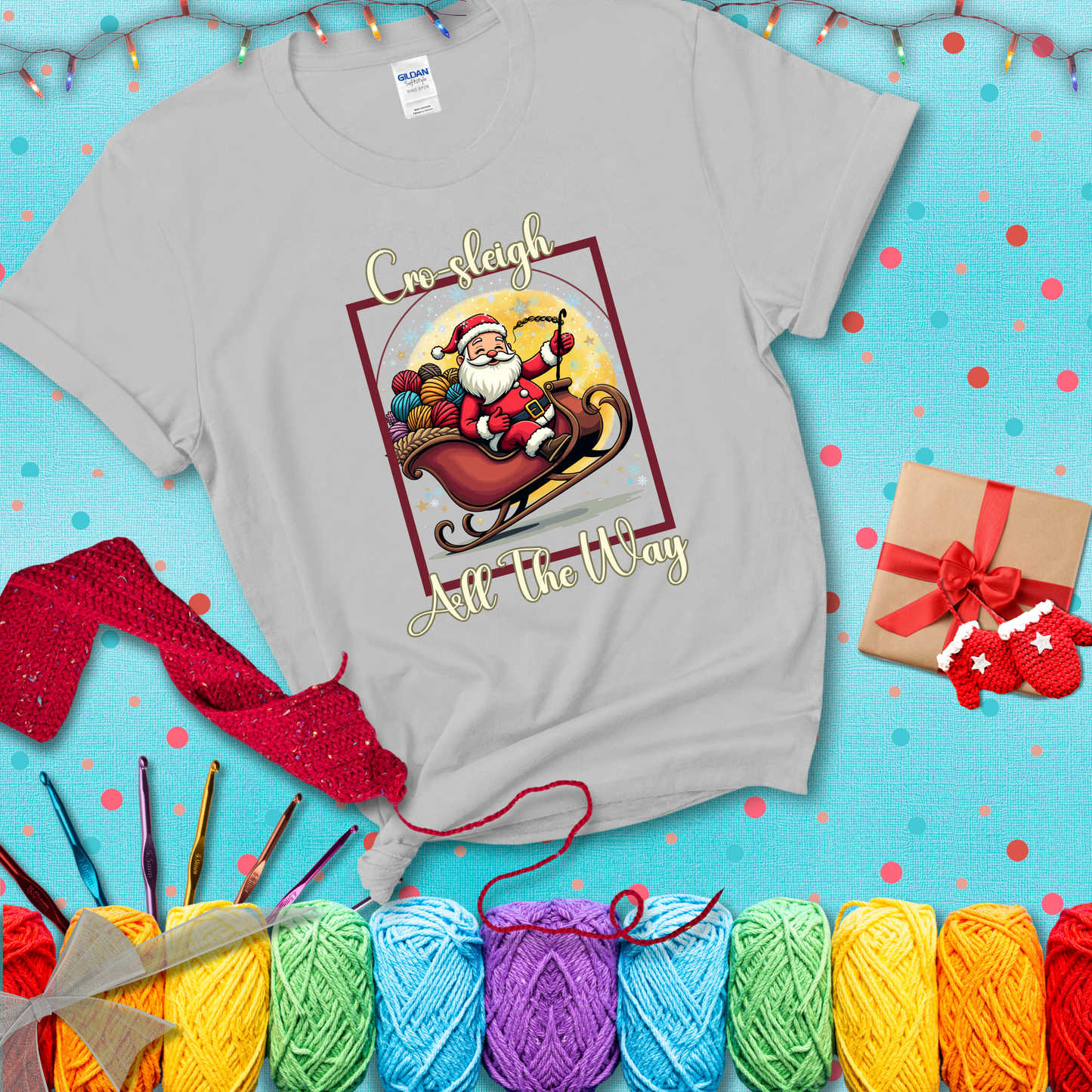 Jolly Santa rides a sleigh overflowing with colorful yarn balls, surrounded by sparkles and holiday cheer. Text reads “Cro-sleigh All the Way" showing a light grey tee with a festive Christmas background