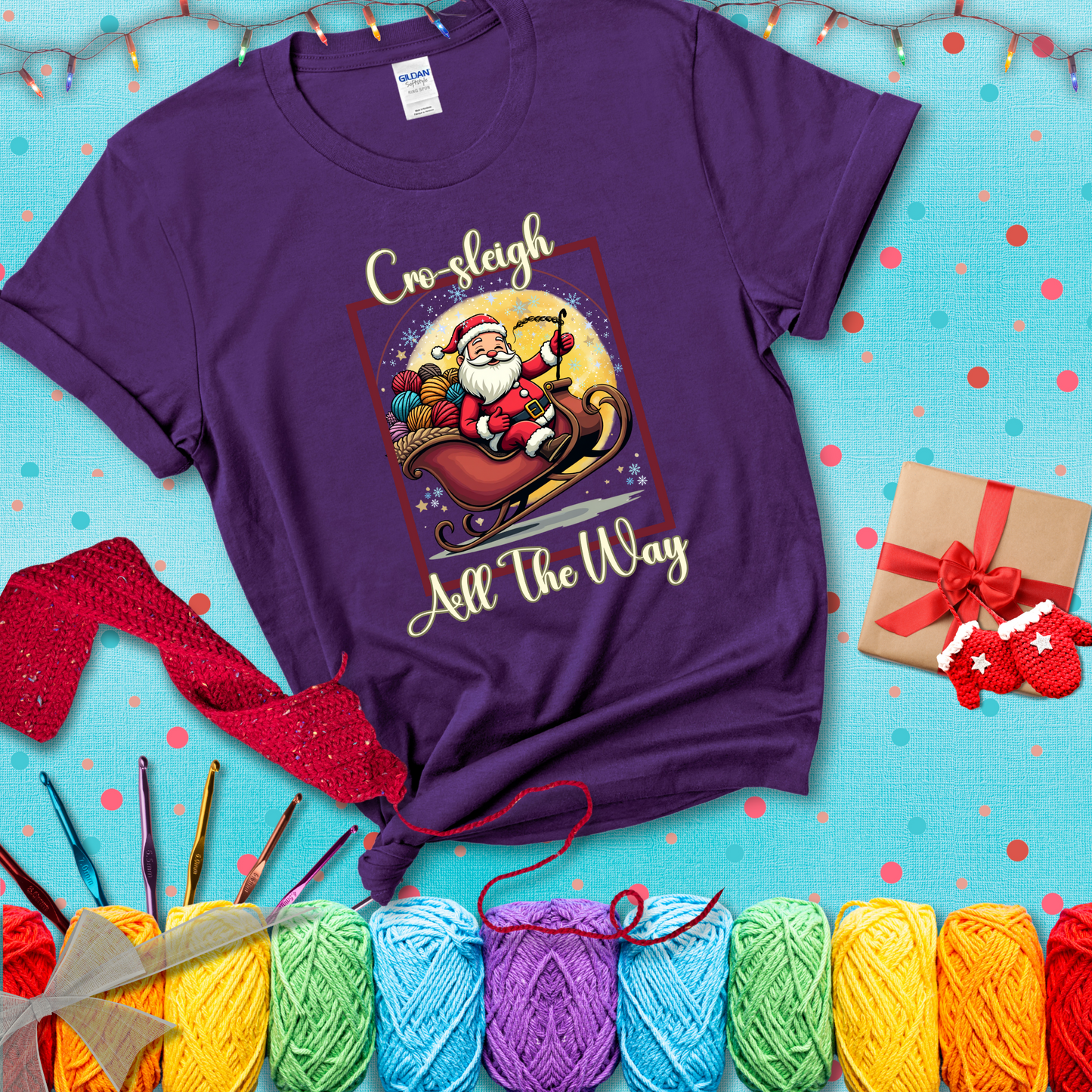 Jolly Santa rides a sleigh overflowing with colorful yarn balls, surrounded by sparkles and holiday cheer. Text reads “Cro-sleigh All the Way" showing a purple tee with a festive Christmas background