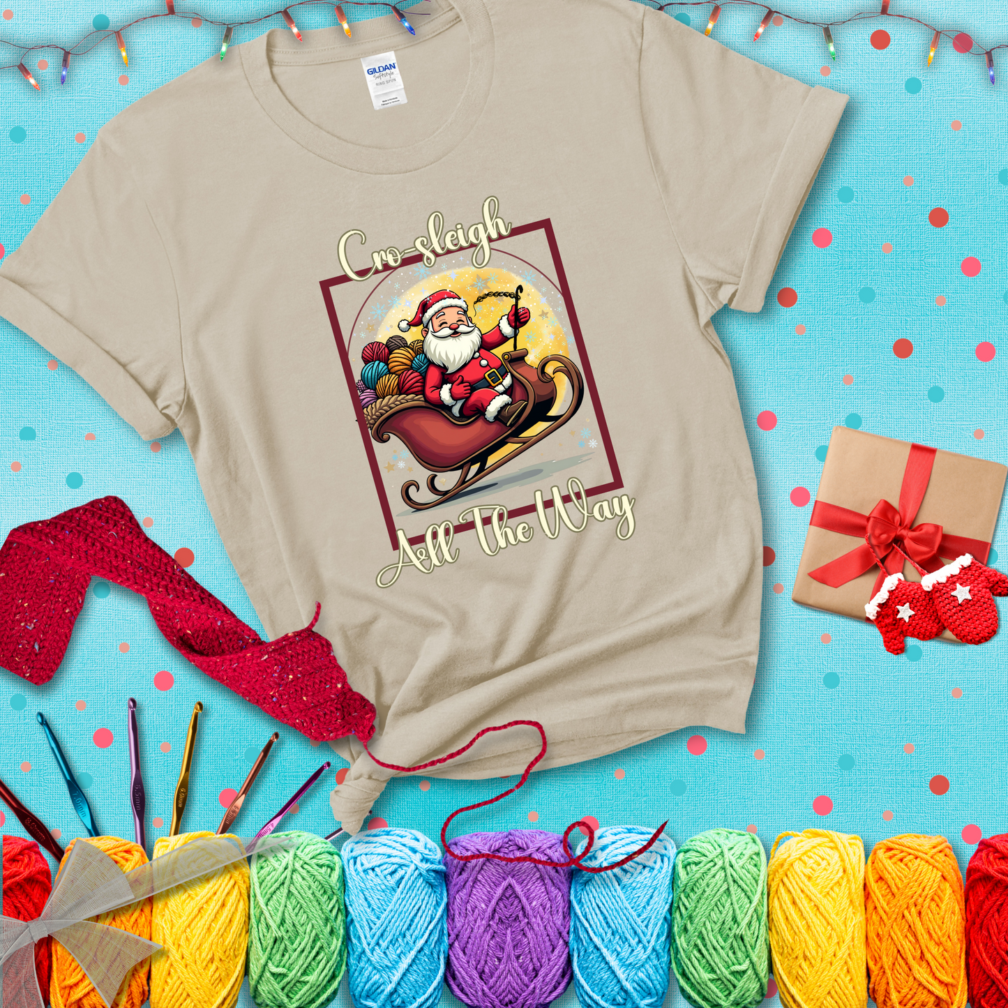 Jolly Santa rides a sleigh overflowing with colorful yarn balls, surrounded by sparkles and holiday cheer. Text reads “Cro-sleigh All the Way" showing a beige tee with a festive Christmas background