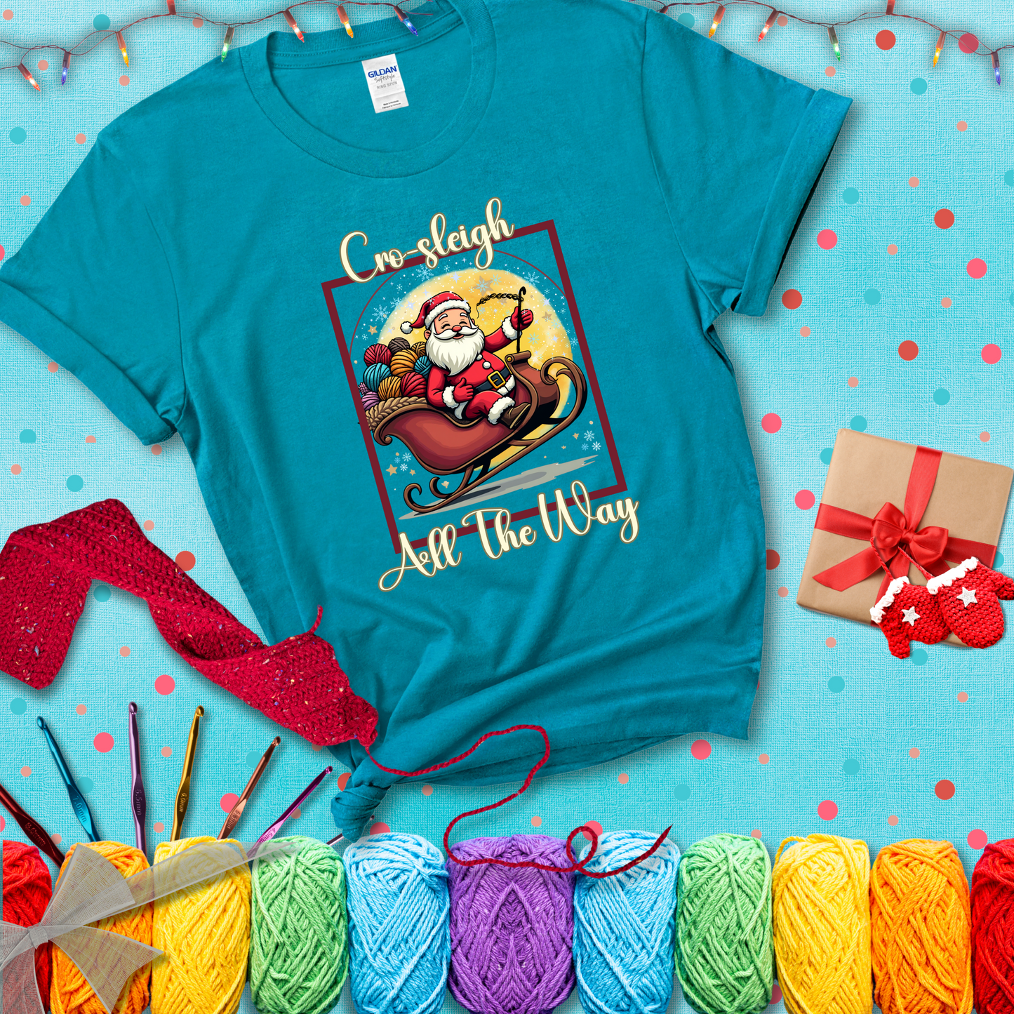 Jolly Santa rides a sleigh overflowing with colorful yarn balls, surrounded by sparkles and holiday cheer. Text reads “Cro-sleigh All the Way" showing a blue-green tee with a festive Christmas background
