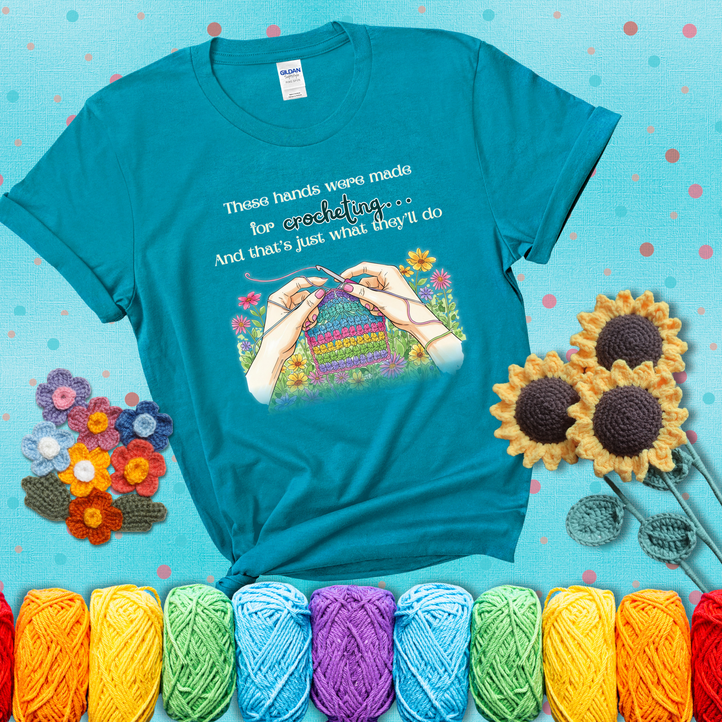Illustration of two hands crocheting a rainbow swatch, surrounded by vibrant flowers with the phrase “These hands were made for crocheting… and that’s just what they’ll do,” showing a blue-green tee with a teal sunflower background