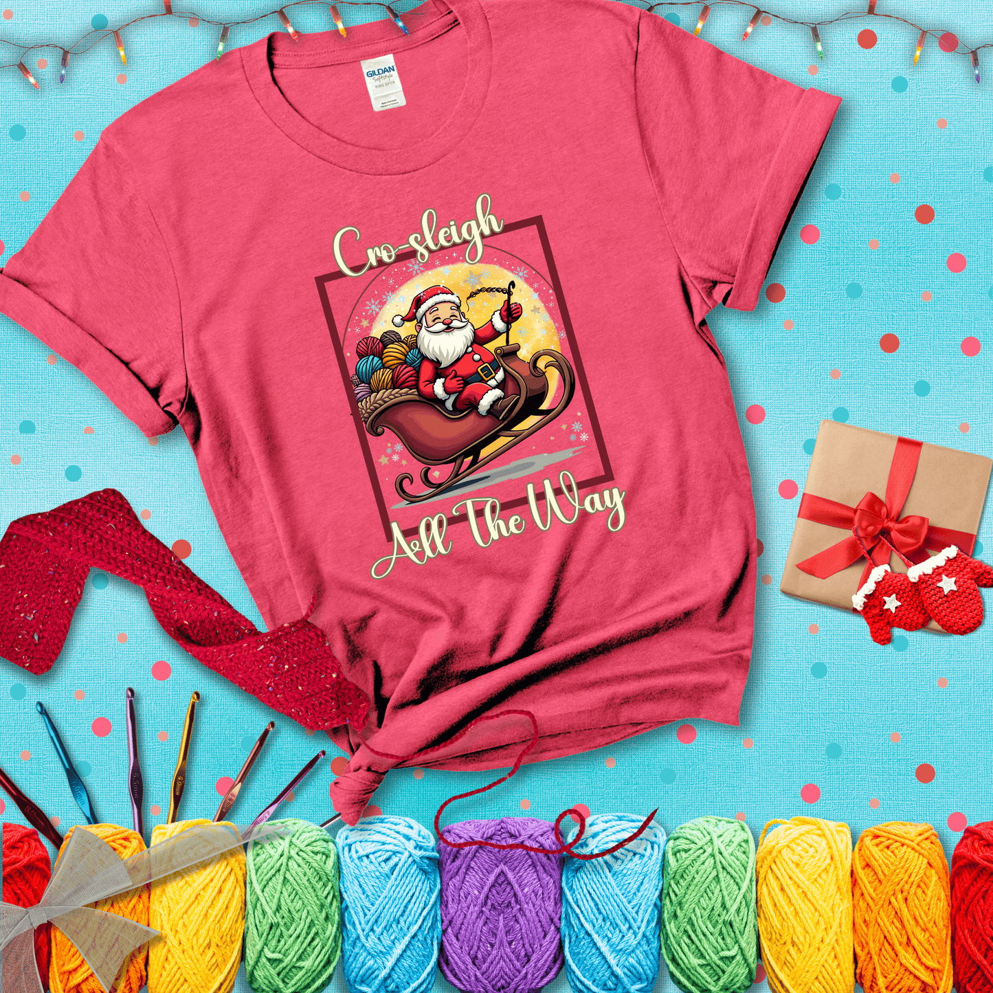 Jolly Santa rides a sleigh overflowing with colorful yarn balls, surrounded by sparkles and holiday cheer. Text reads “Cro-sleigh All the Way" showing a light red tee with a festive Christmas background