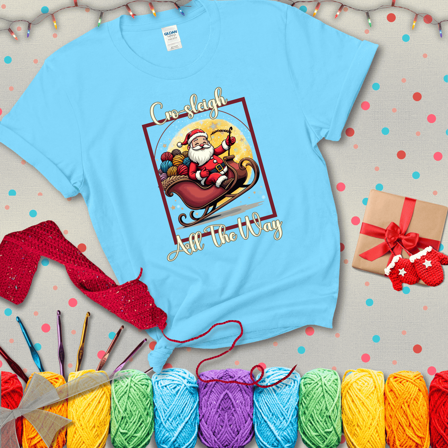 Jolly Santa rides a sleigh overflowing with colorful yarn balls, surrounded by sparkles and holiday cheer. Text reads “Cro-sleigh All the Way" showing a light blue tee with a festive Christmas background
