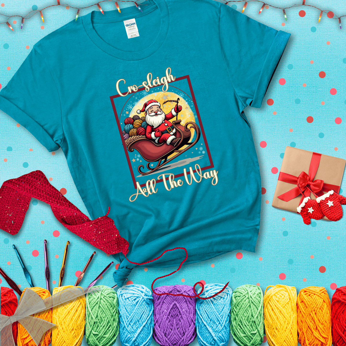 Jolly Santa rides a sleigh overflowing with colorful yarn balls, surrounded by sparkles and holiday cheer. Text reads “Cro-sleigh All the Way" showing a blue-green tee with a festive Christmas background