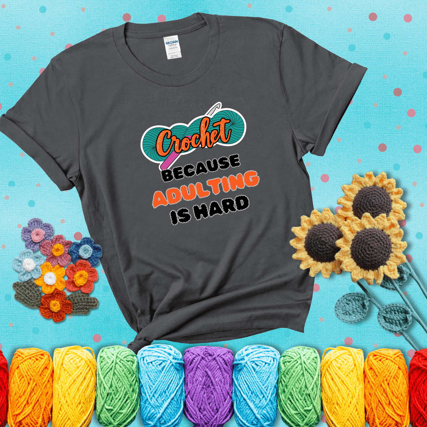 A bold design with the phrase “Crochet Because Adulting Is Hard” featuring a green skein of yarn with a pink crochet hook. The word “Crochet” is in a script font, while “Adulting is hard” is in chunky textured letters showing a dark grey tee with a teal sunflower background