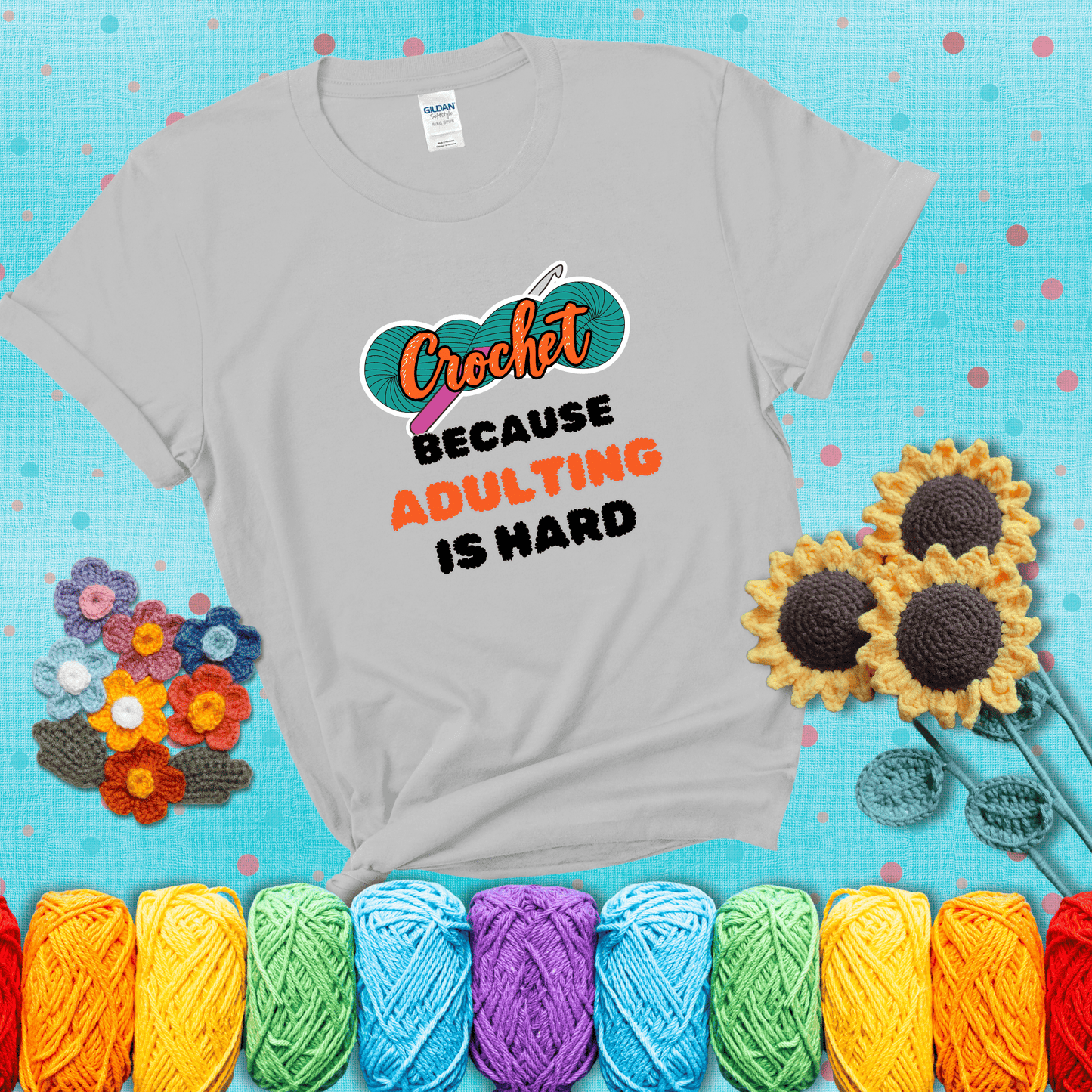 A bold design with the phrase “Crochet Because Adulting Is Hard” featuring a green skein of yarn with a pink crochet hook. The word “Crochet” is in a script font, while “Adulting is hard” is in chunky textured letters showing a light grey tee with a teal sunflower background
