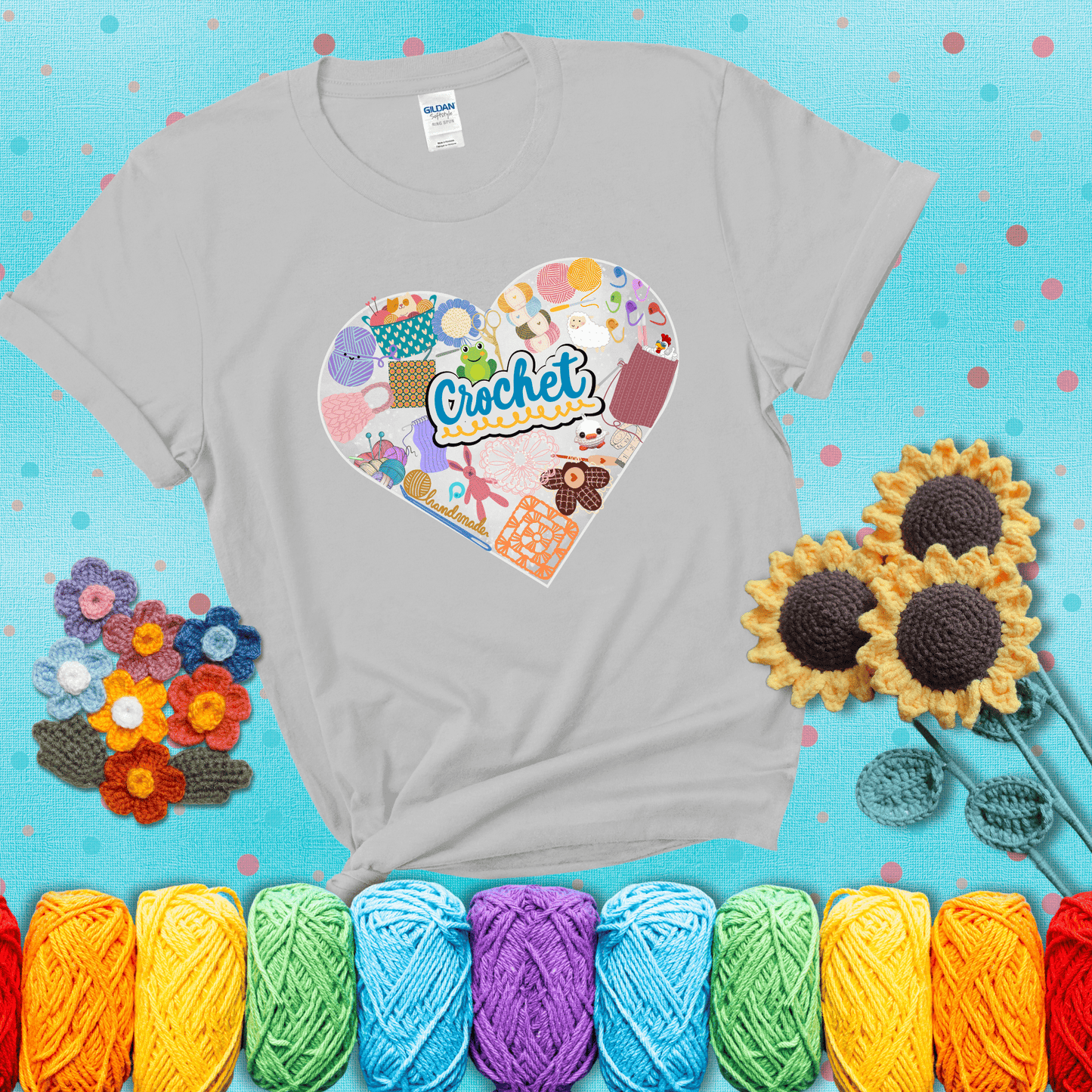A heart filled with colorful crochet essentials including yarn, hooks, scissors, notions, and handmade items, with the word "Crochet" prominently in the center, showing a light grey tee with a teal sunflower background
