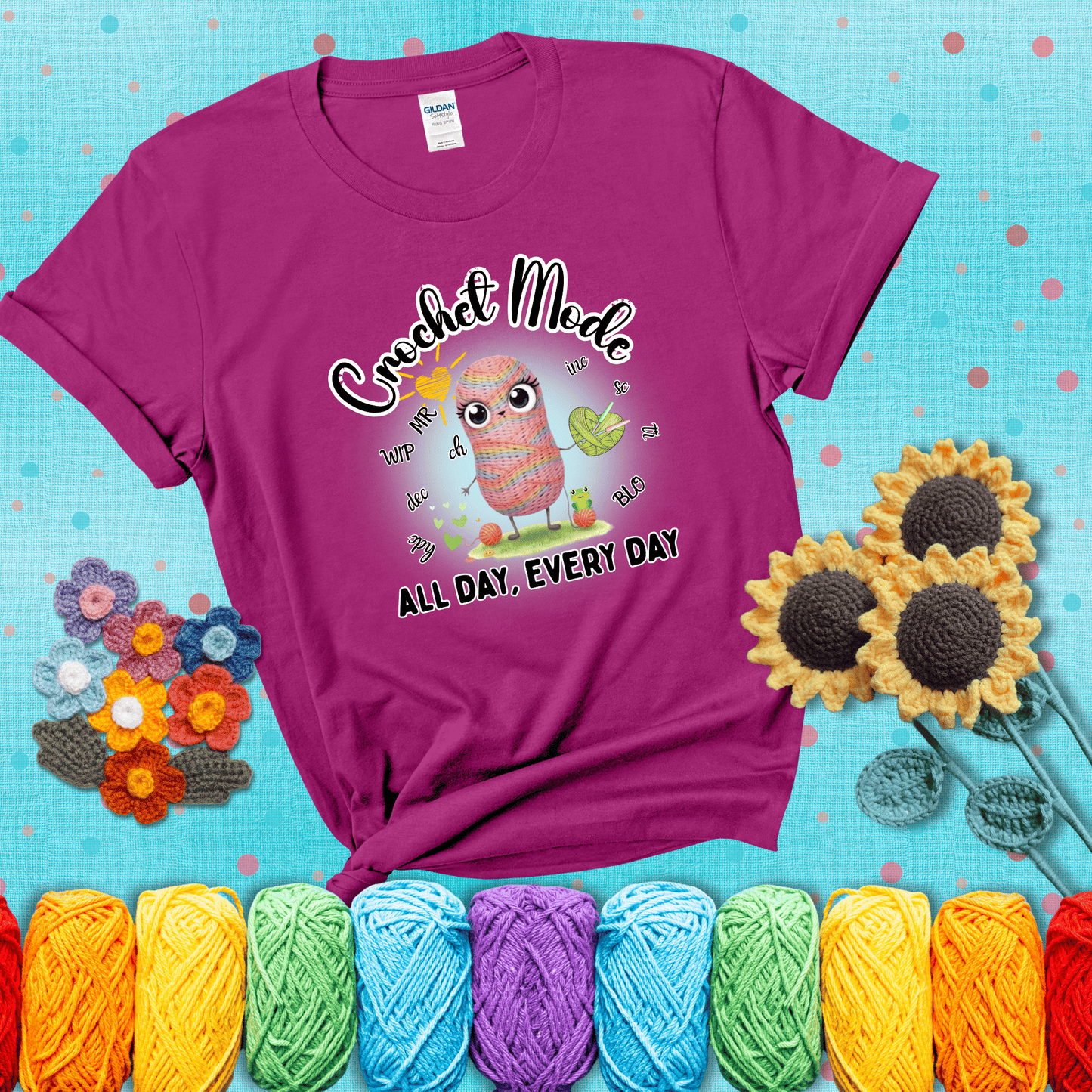 A kawaii-style rainbow yarn character surrounded by crochet stitch abbreviations and holding a yarn heart, with text “Crochet Mode All Day, Every Day"; showing a magenta tee with a teal sunflower background