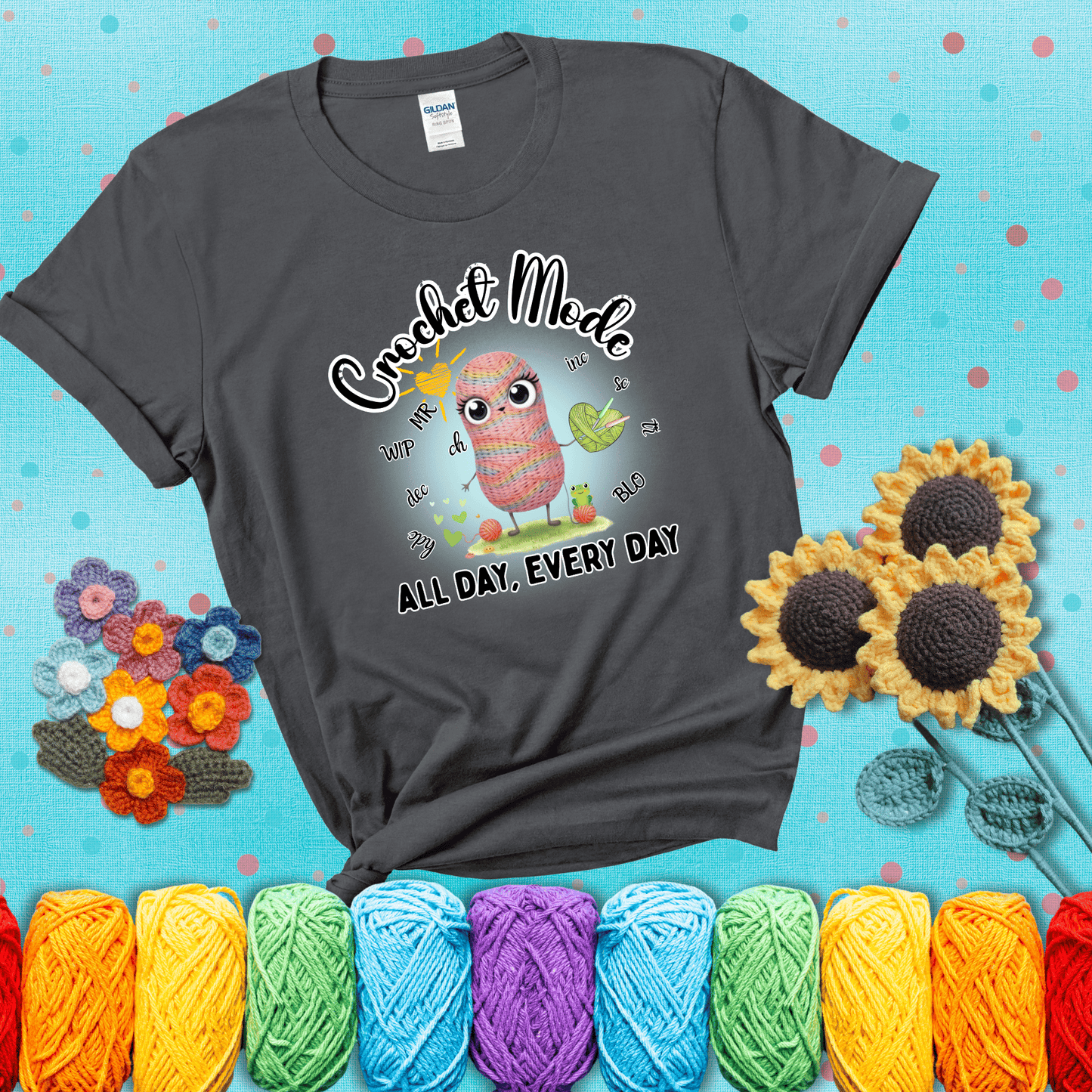 A kawaii-style rainbow yarn character surrounded by crochet stitch abbreviations and holding a yarn heart, with text “Crochet Mode All Day, Every Day"; showing a dark grey tee with a teal sunflower background