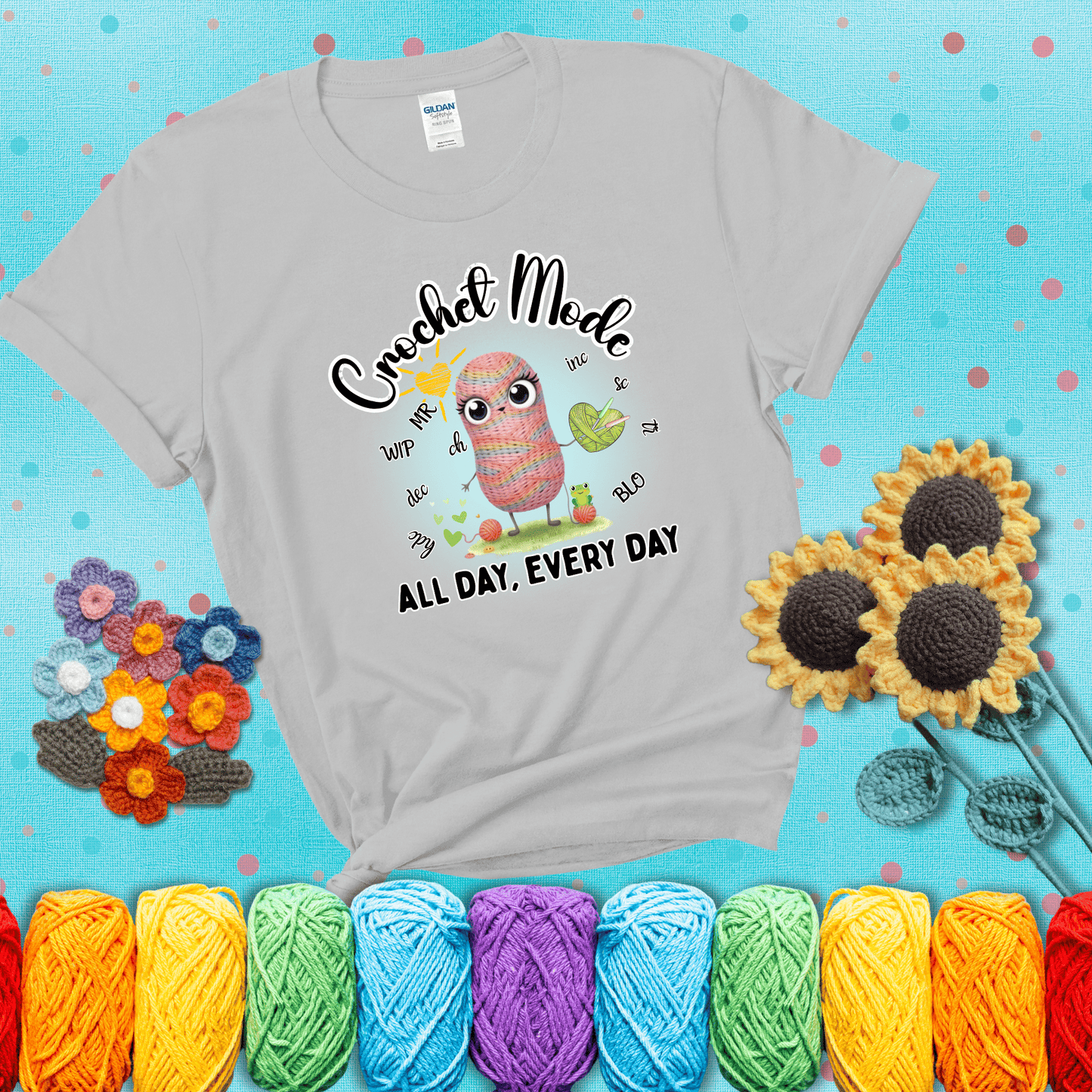 A kawaii-style rainbow yarn character surrounded by crochet stitch abbreviations and holding a yarn heart, with text “Crochet Mode All Day, Every Day"; showing a light grey tee with a teal sunflower background