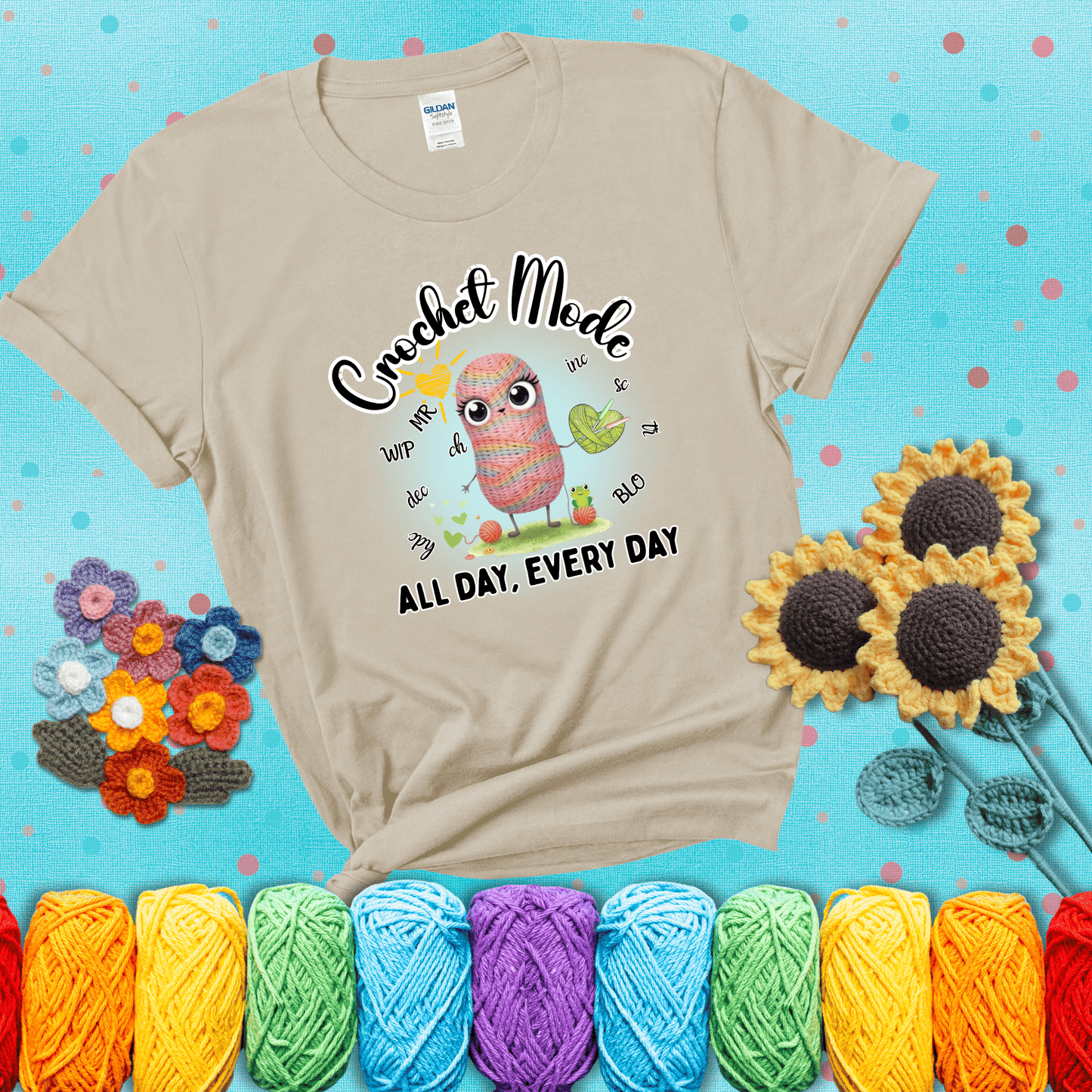 A kawaii-style rainbow yarn character surrounded by crochet stitch abbreviations and holding a yarn heart, with text “Crochet Mode All Day, Every Day"; showing a beige tee with a teal sunflower background